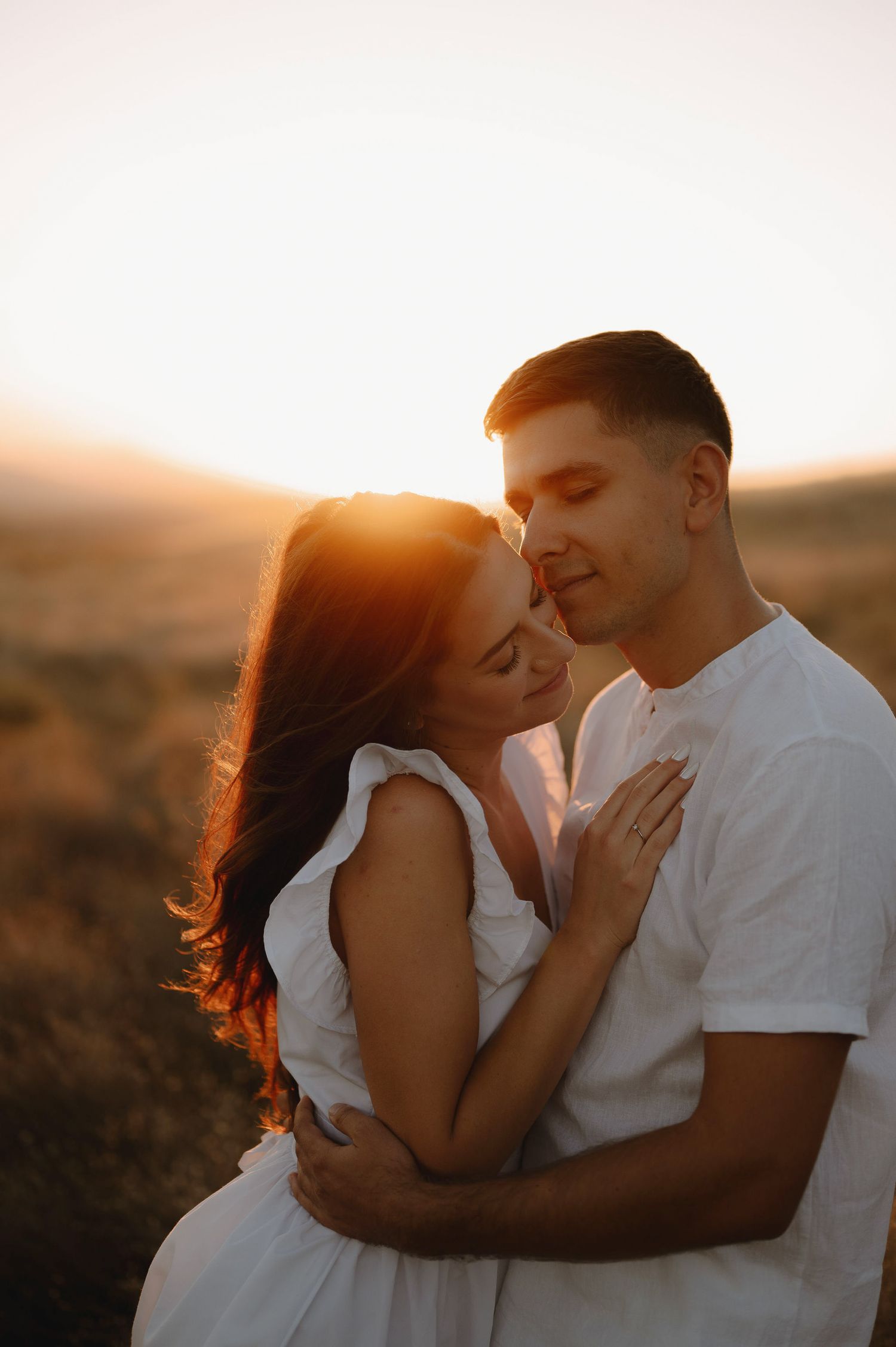 A romantic couple embraces during golden hour in a field as sunlight creates a warm, dreamy atmosphere.
