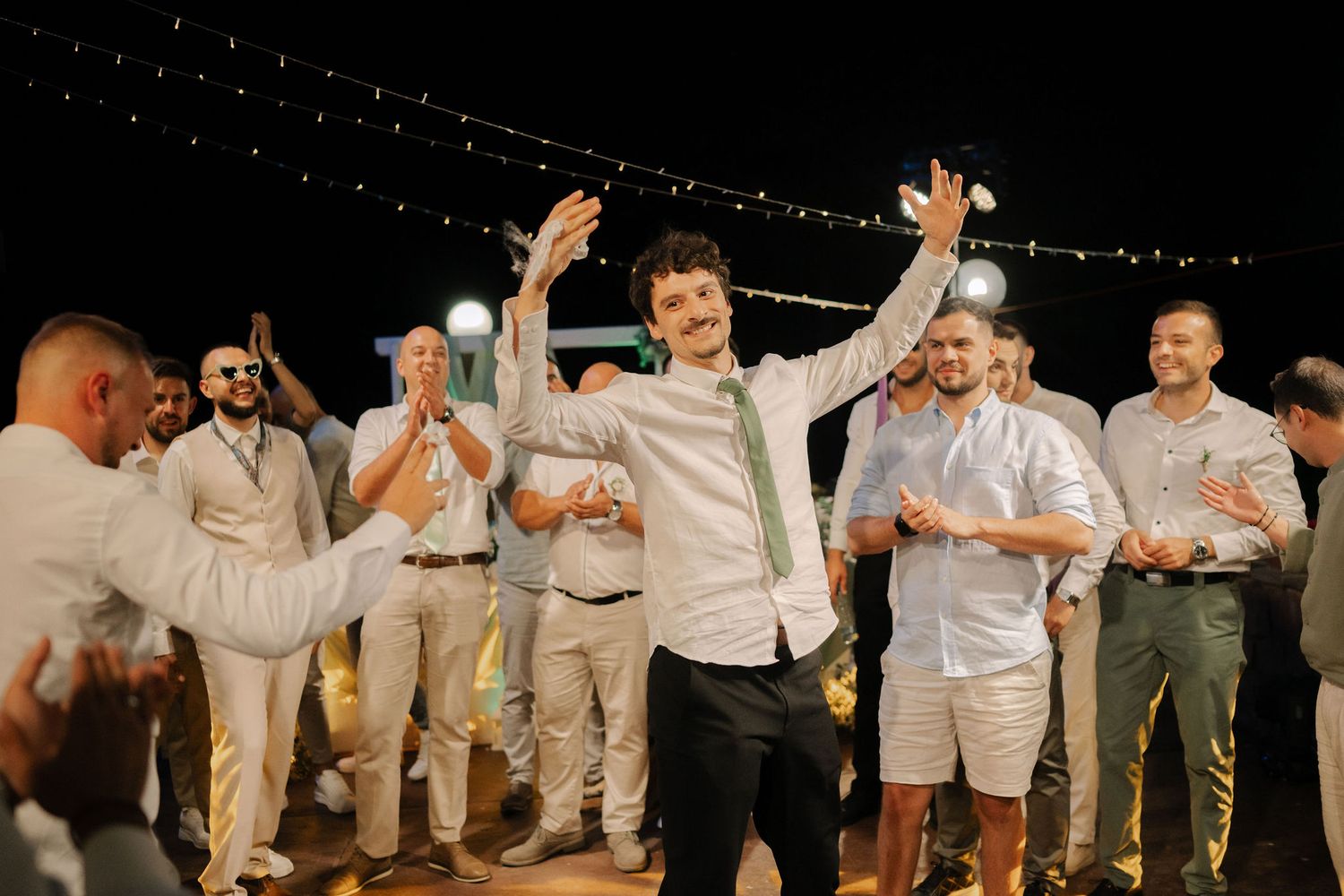 A group of groomsmen in light-colored suits and white shirts dancing in a line at a wedding reception.
