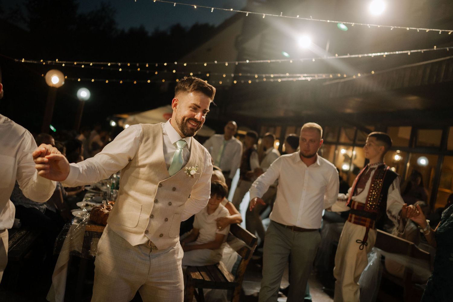 Wedding guests dance and celebrate on an outdoor patio decorated with string lights during an evening reception.
