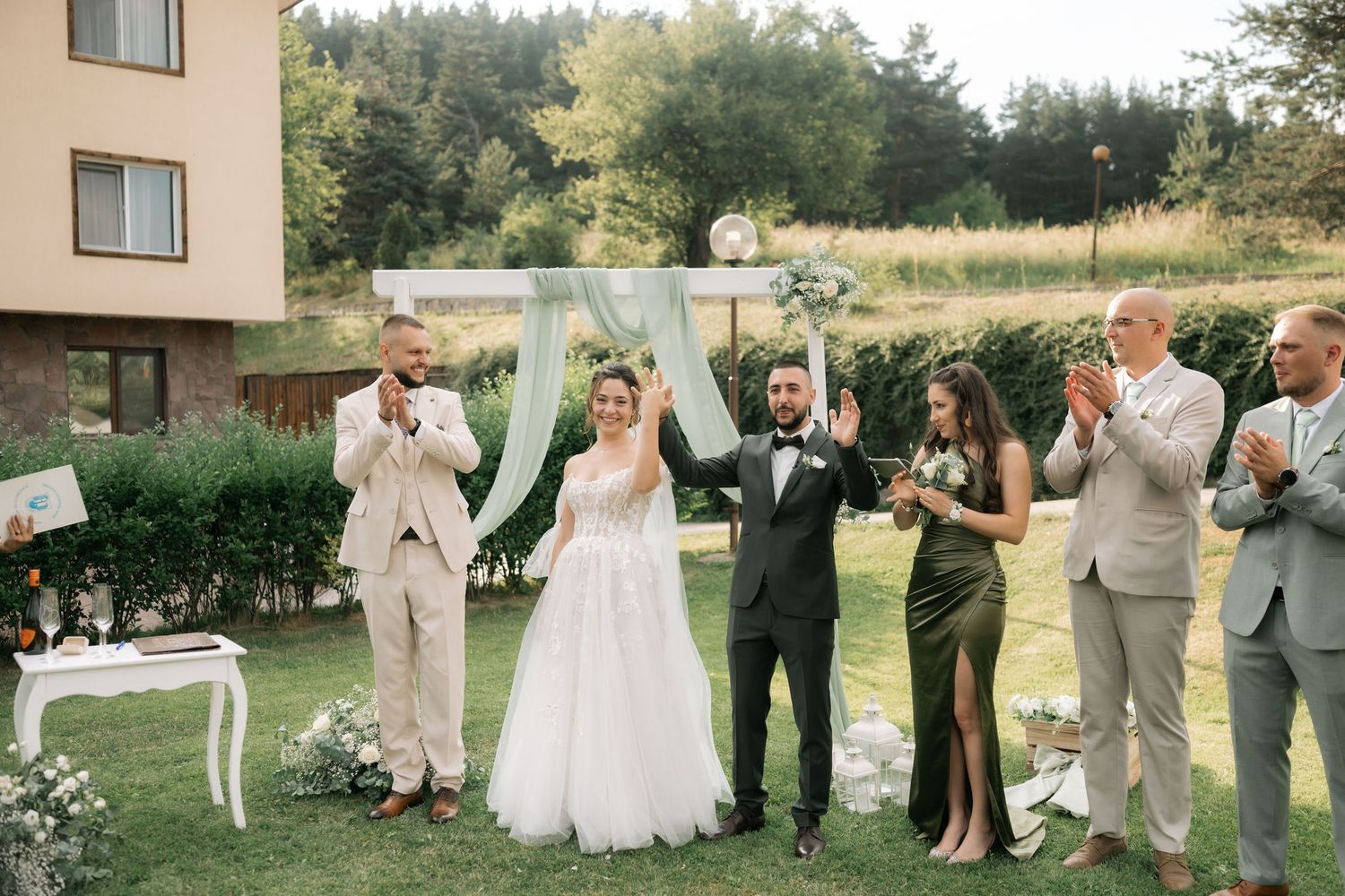 Wedding party members in light colored attire pose together on a grassy lawn during an outdoor summer celebration.