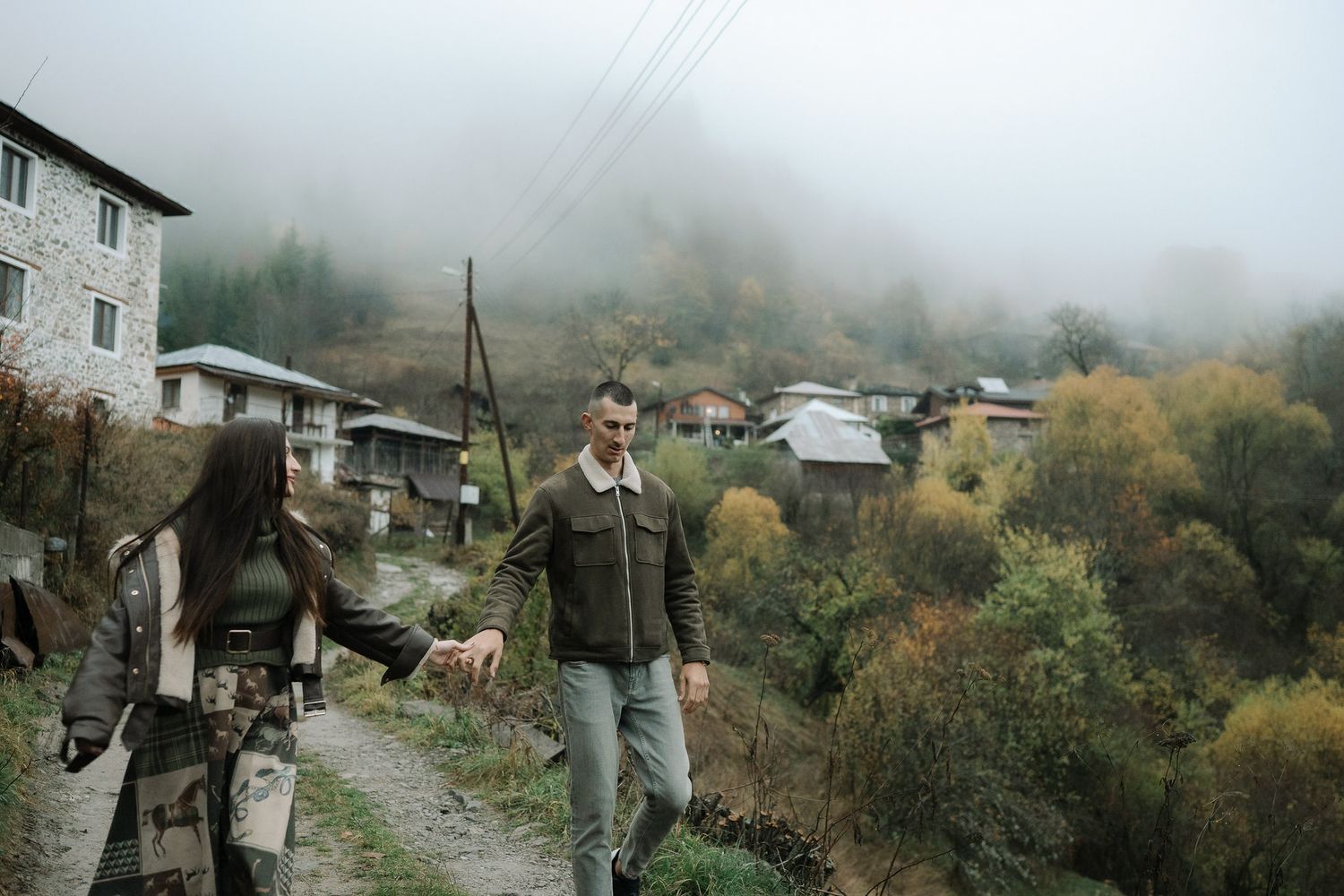 A foggy mountain village path winds through traditional stone houses and autumn trees in a remote rural setting.