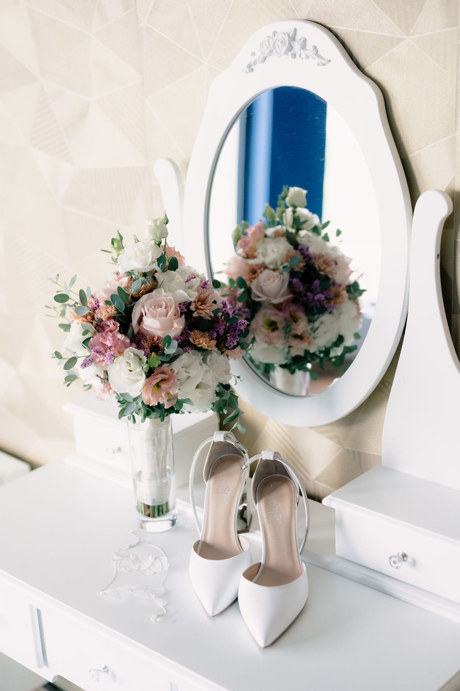 Elegant bridal shoes and floral bouquet with pink roses displayed on white vanity with round mirror.