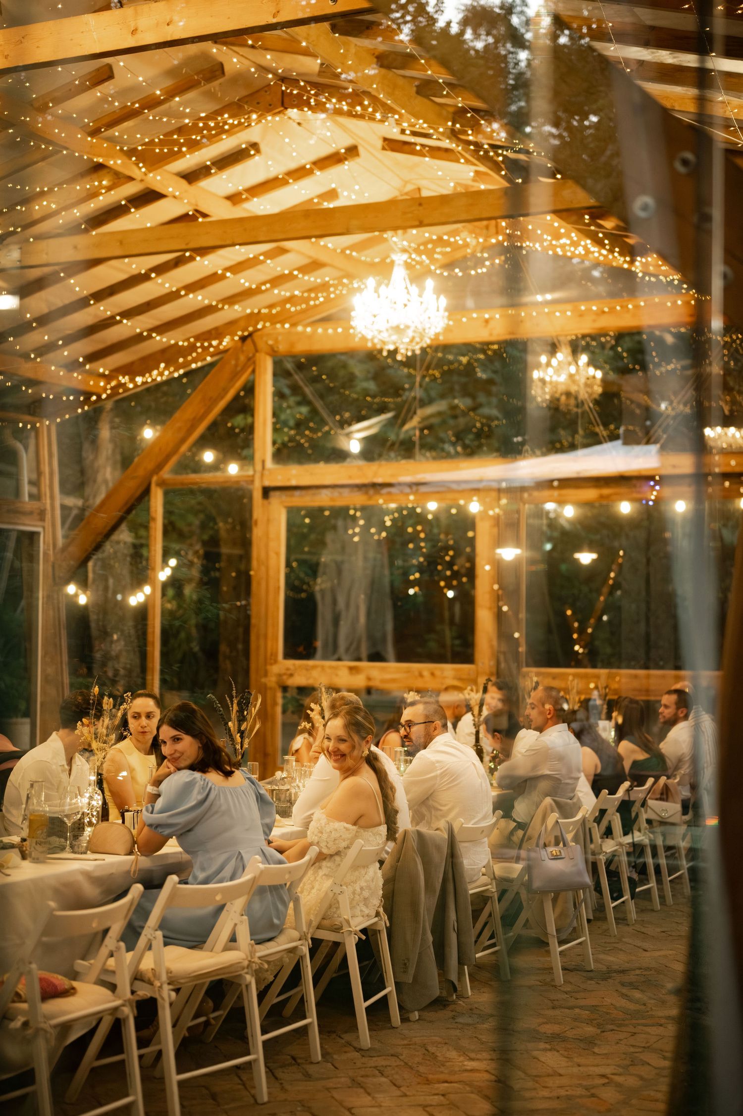 Wedding reception guests dine under a rustic wooden pavilion decorated with twinkling string lights and elegant chandeliers.
