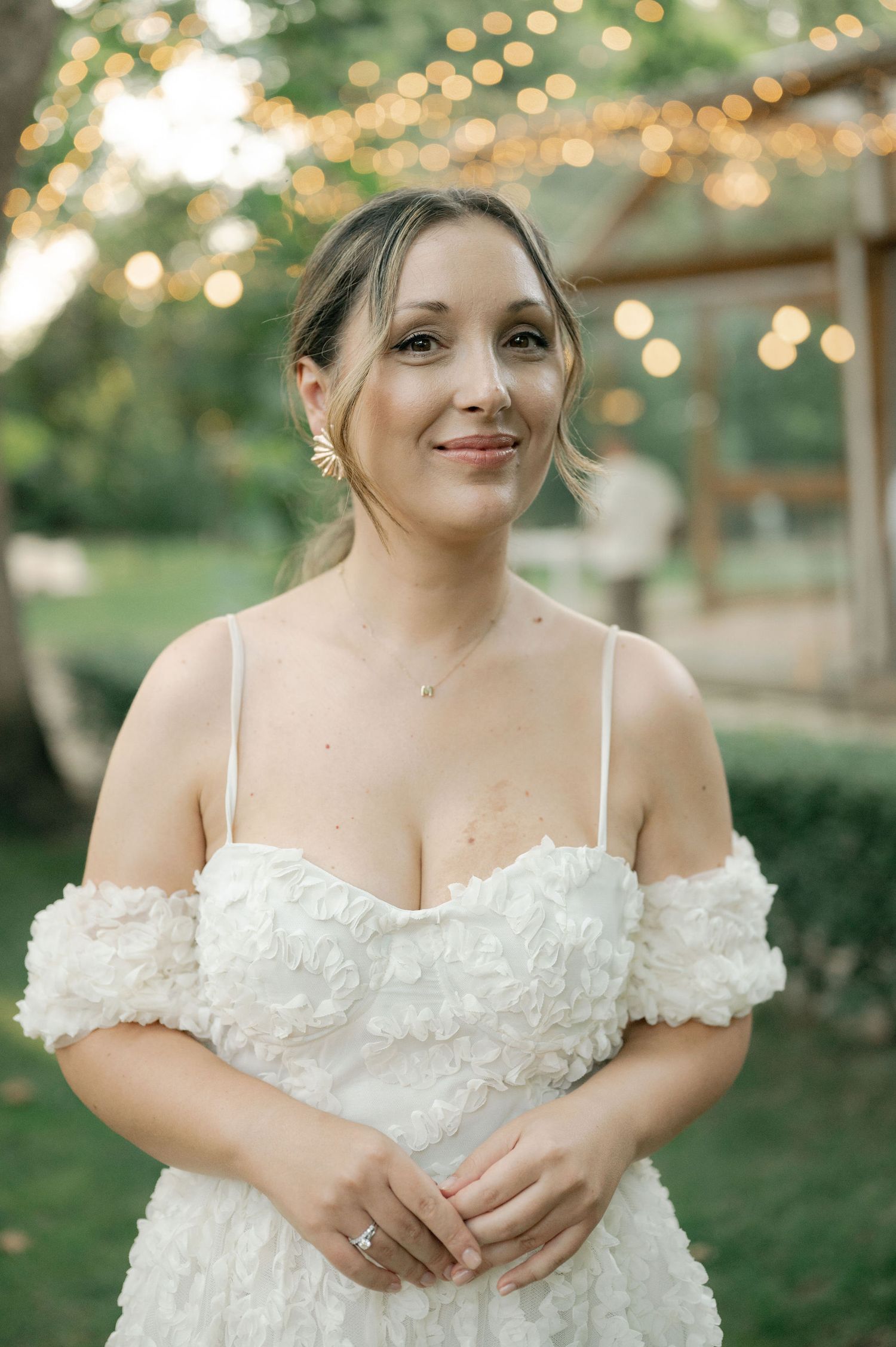 A bride in an off-shoulder white wedding dress with delicate floral details poses in a garden with twinkling lights behind.