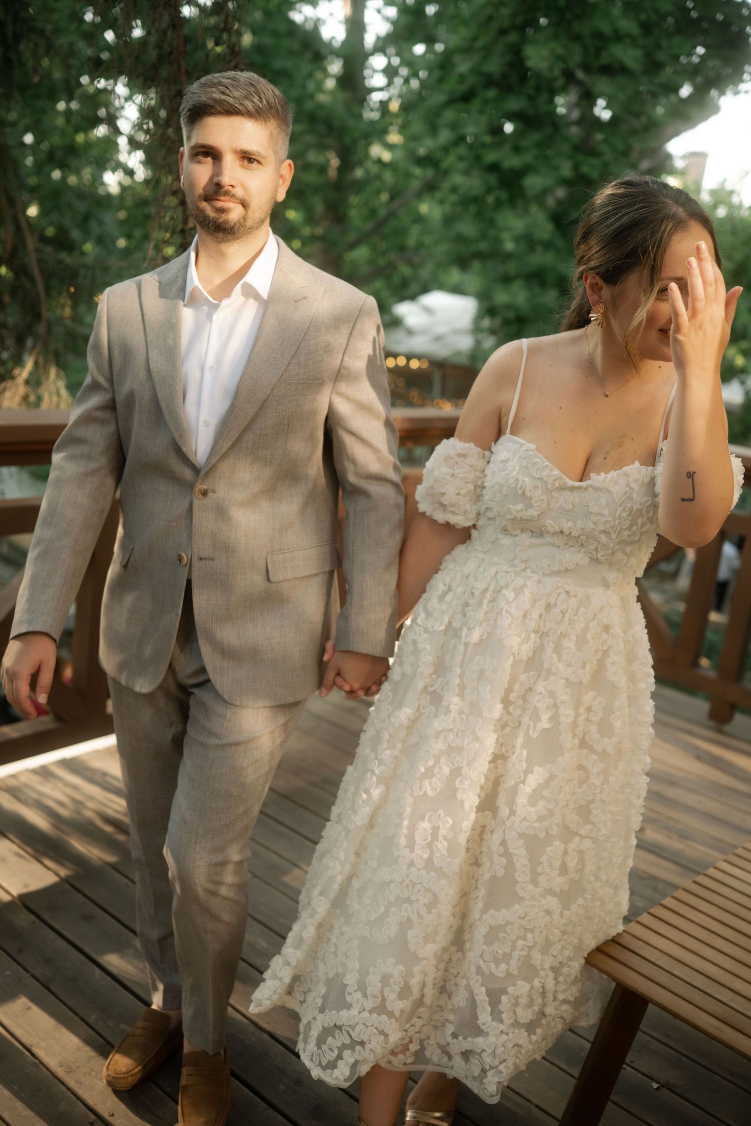 A couple in wedding attire holds hands while walking on a wooden deck surrounded by trees.