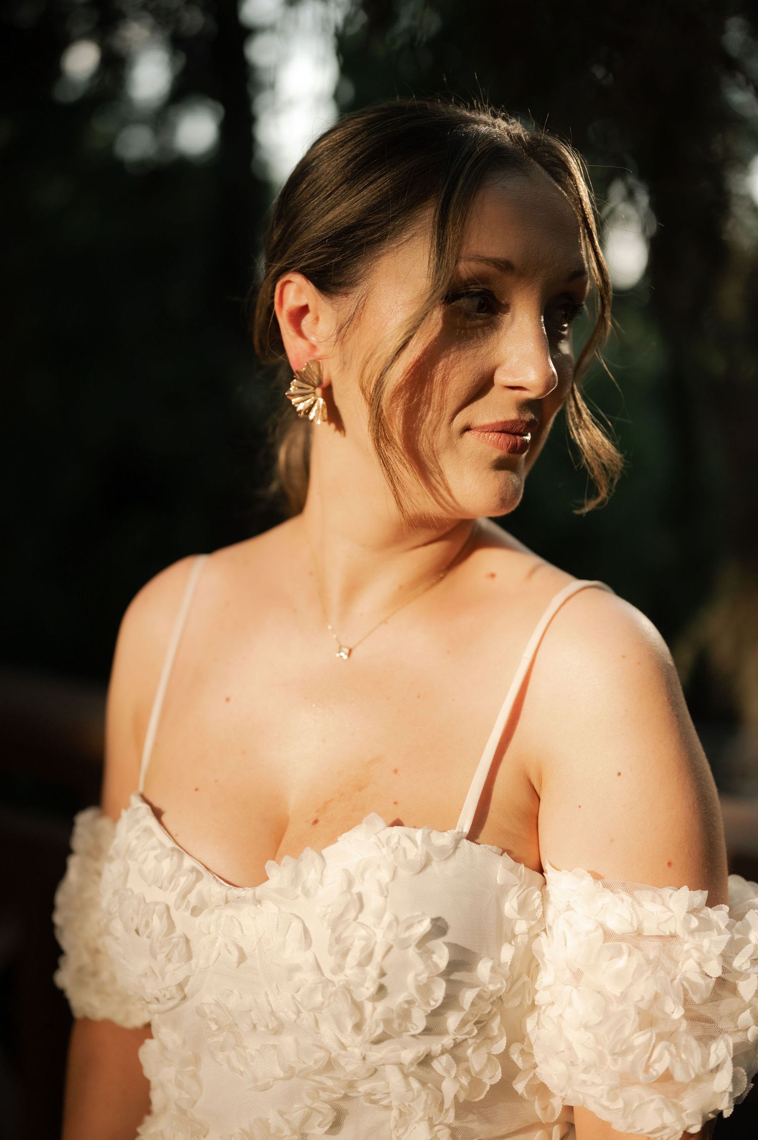 A bride in an off-shoulder wedding dress with delicate floral details poses in a romantic outdoor setting at sunset.