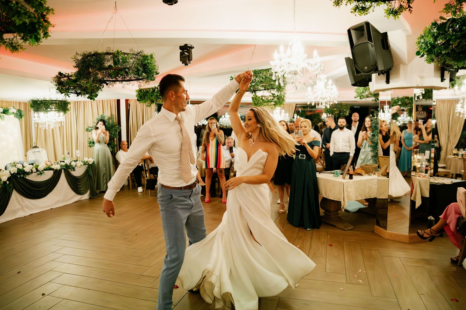 Wedding guests enjoying themselves at a lively dance party in an elegant reception hall with greenery decor.