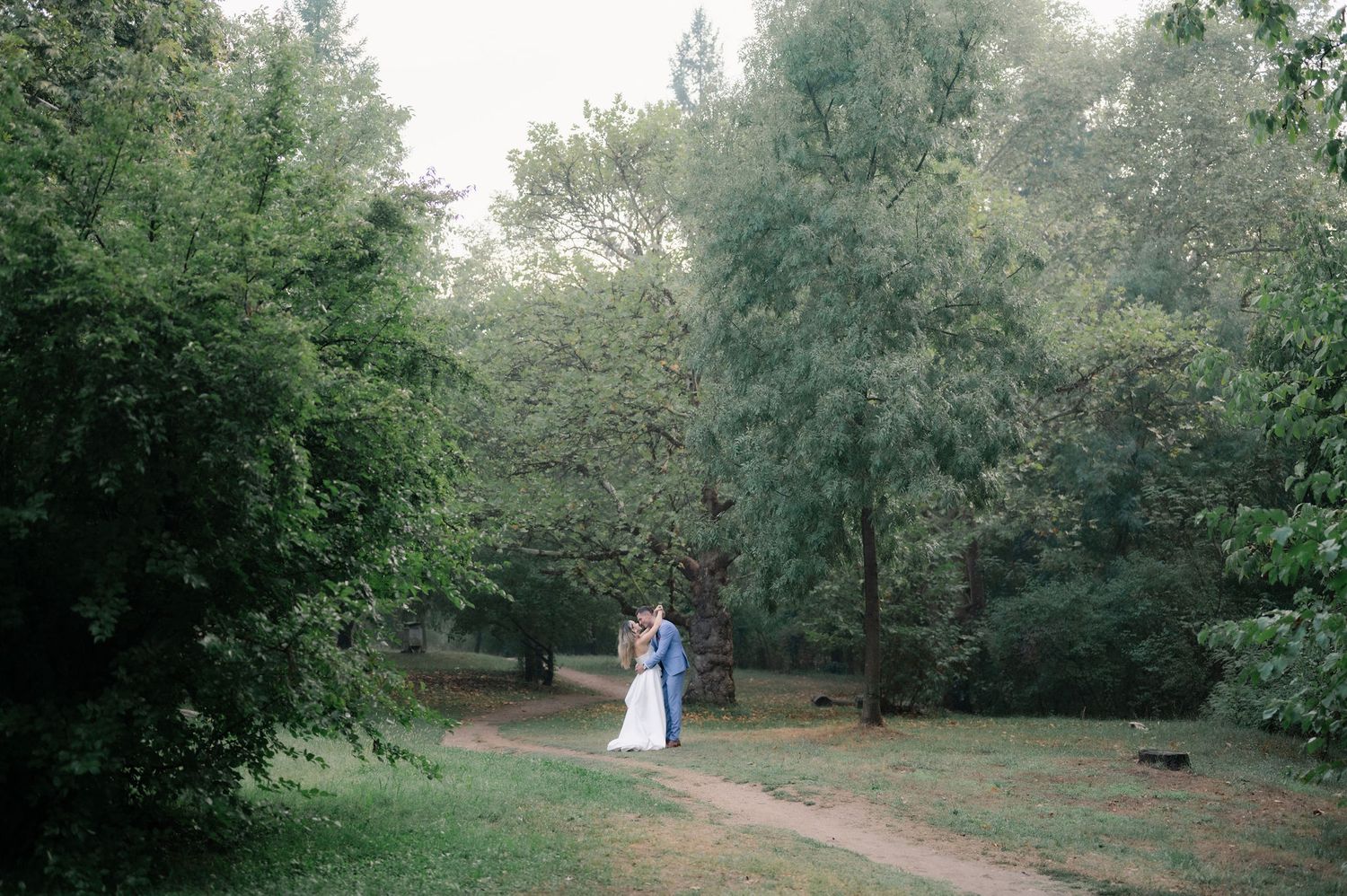 Two people in formal attire walk along a serene wooded path surrounded by lush green trees.