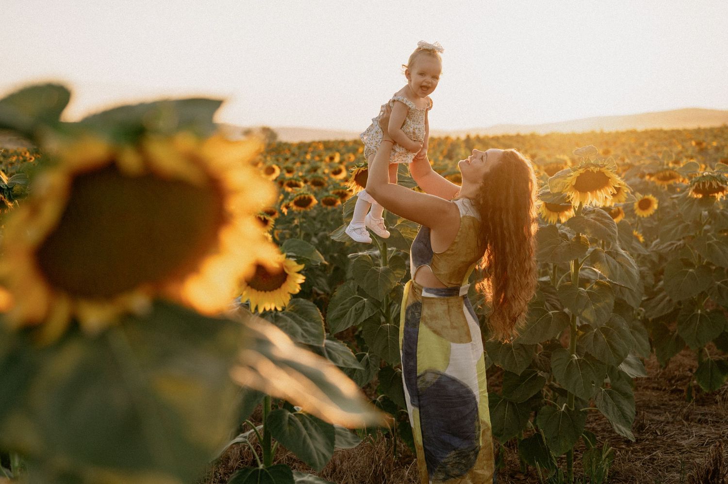 A mother lifts her toddler joyfully in a golden sunflower field at sunset.
