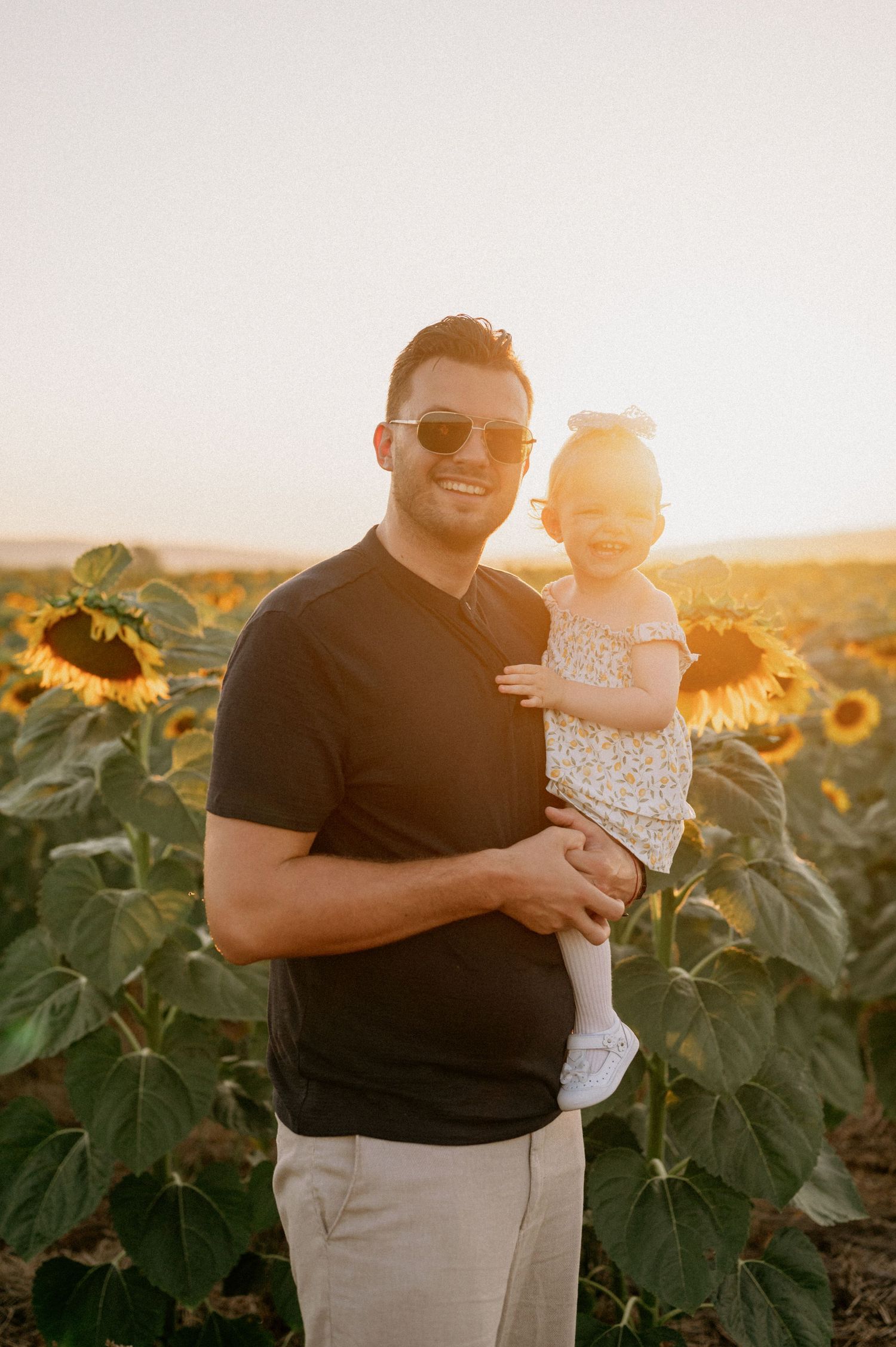 Man in sunglasses holds smiling toddler girl in sunflower field during golden hour sunset.