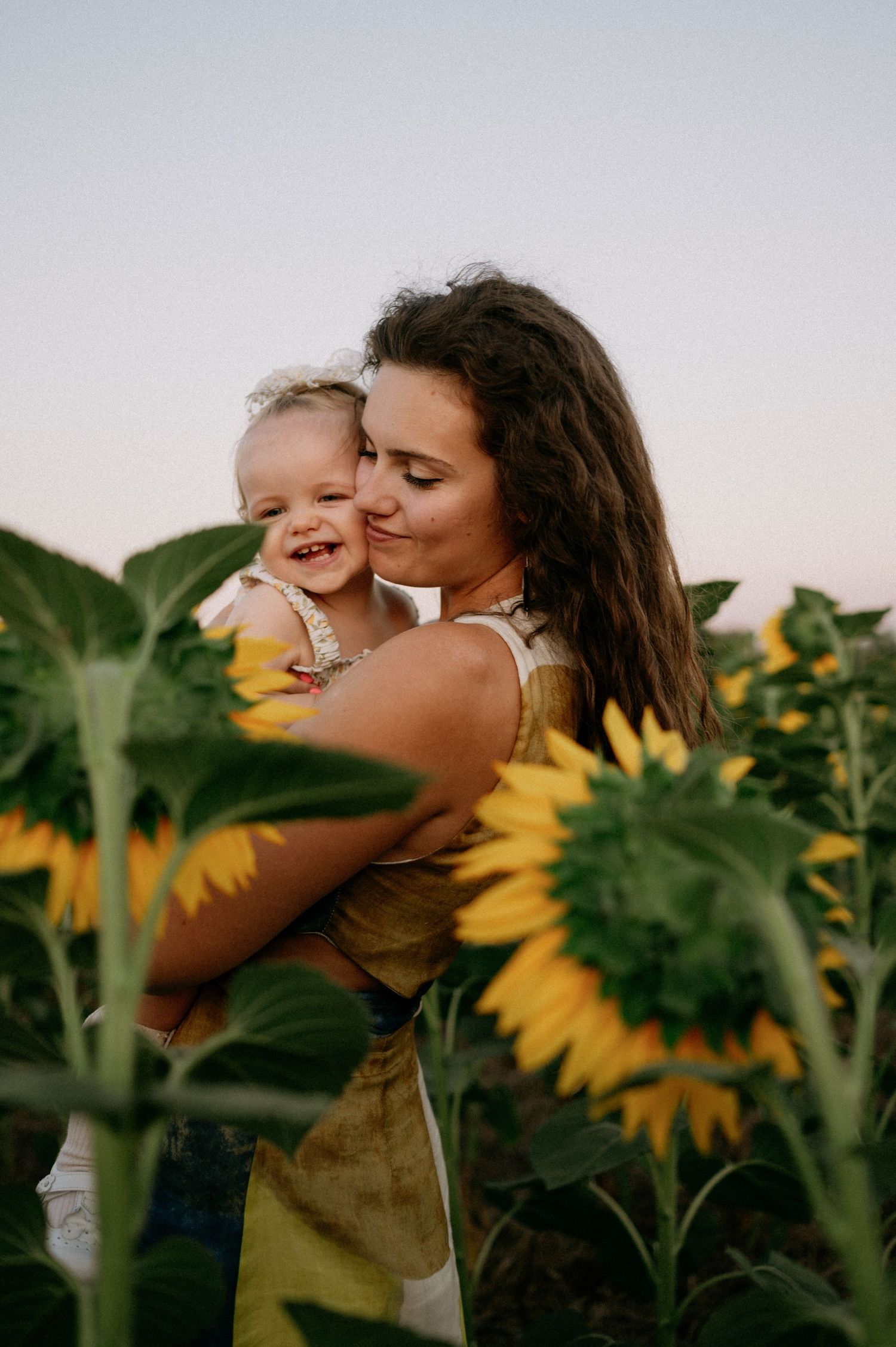 A young mother kisses her laughing baby girl in a vibrant sunflower field at dusk.