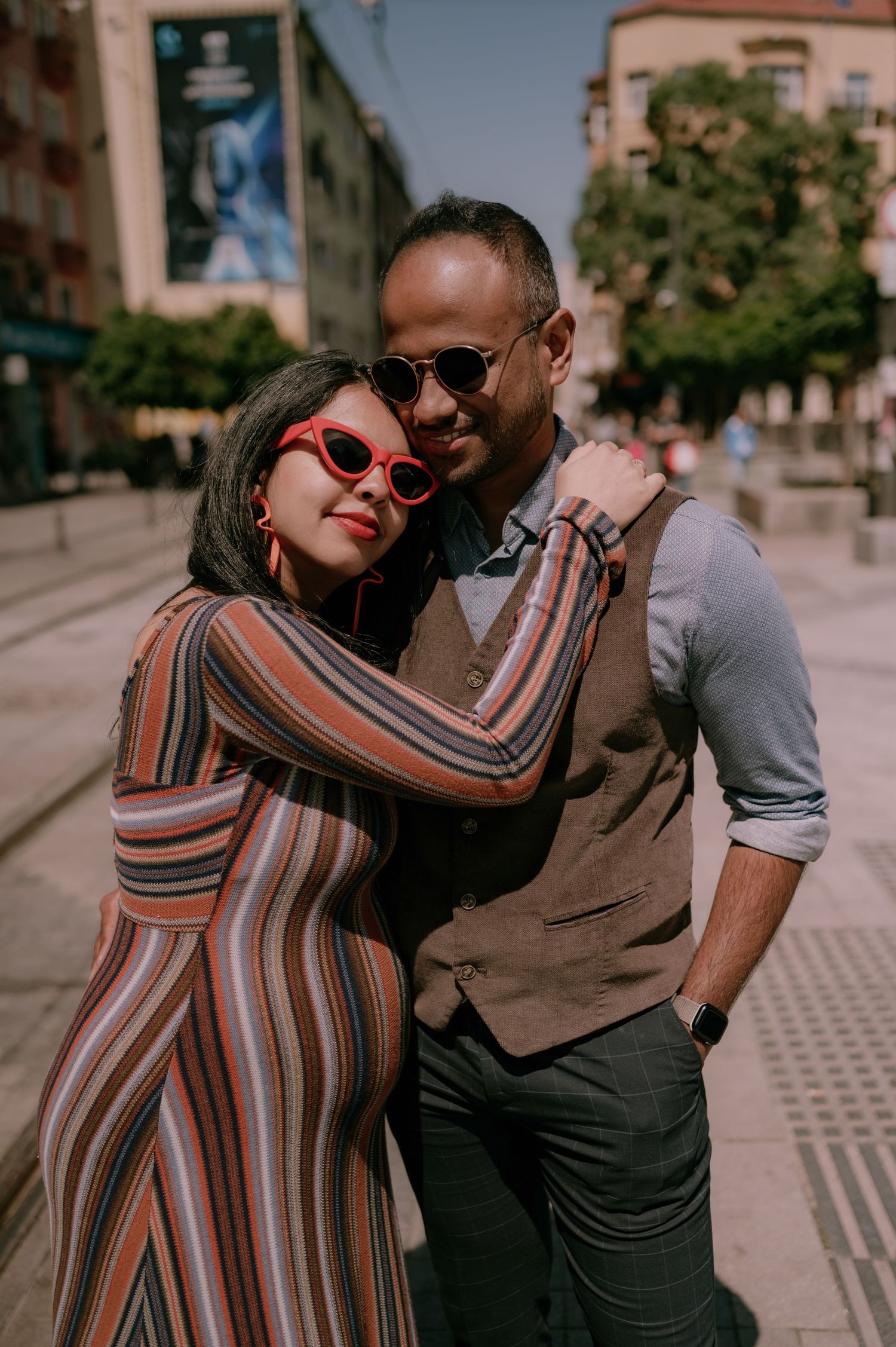 Stylish couple embracing on a sunny city street, woman in striped dress and red sunglasses, man in vest and round shades.