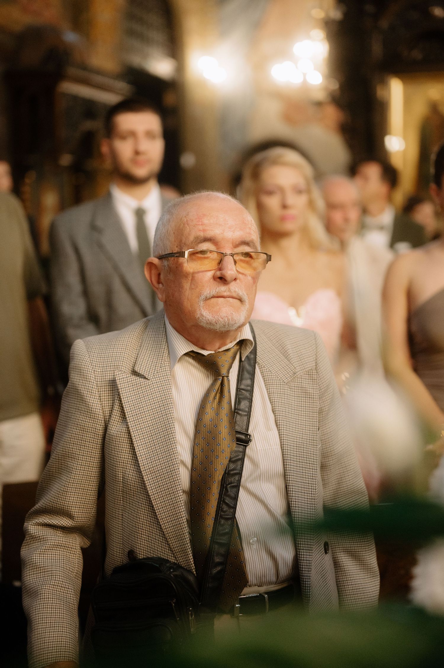 An elderly man in a beige suit and glasses sits attentively at what appears to be a formal event or gathering.