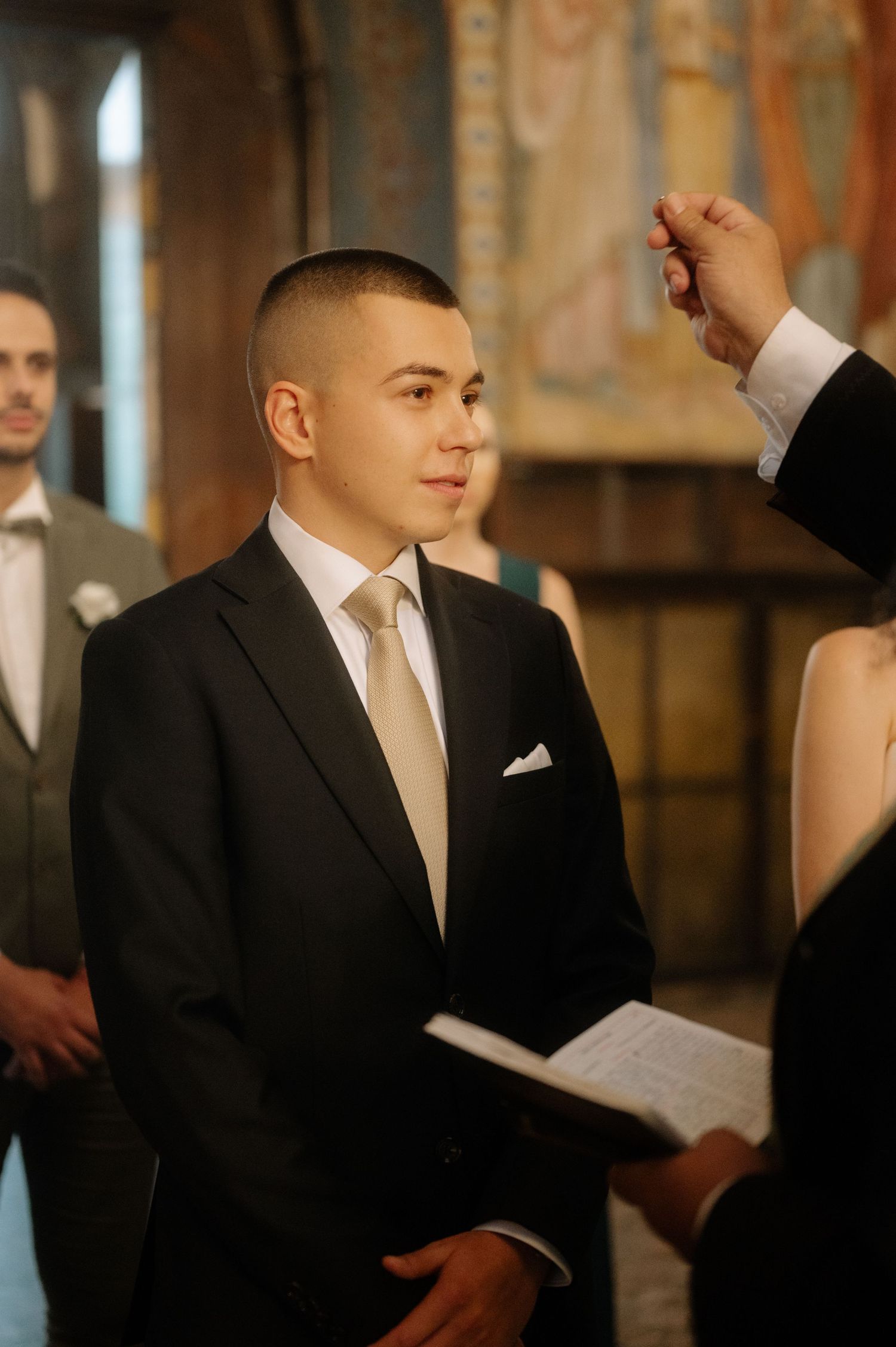 A young groom in a black suit and light tie stands attentively during a wedding ceremony in an ornate church setting.