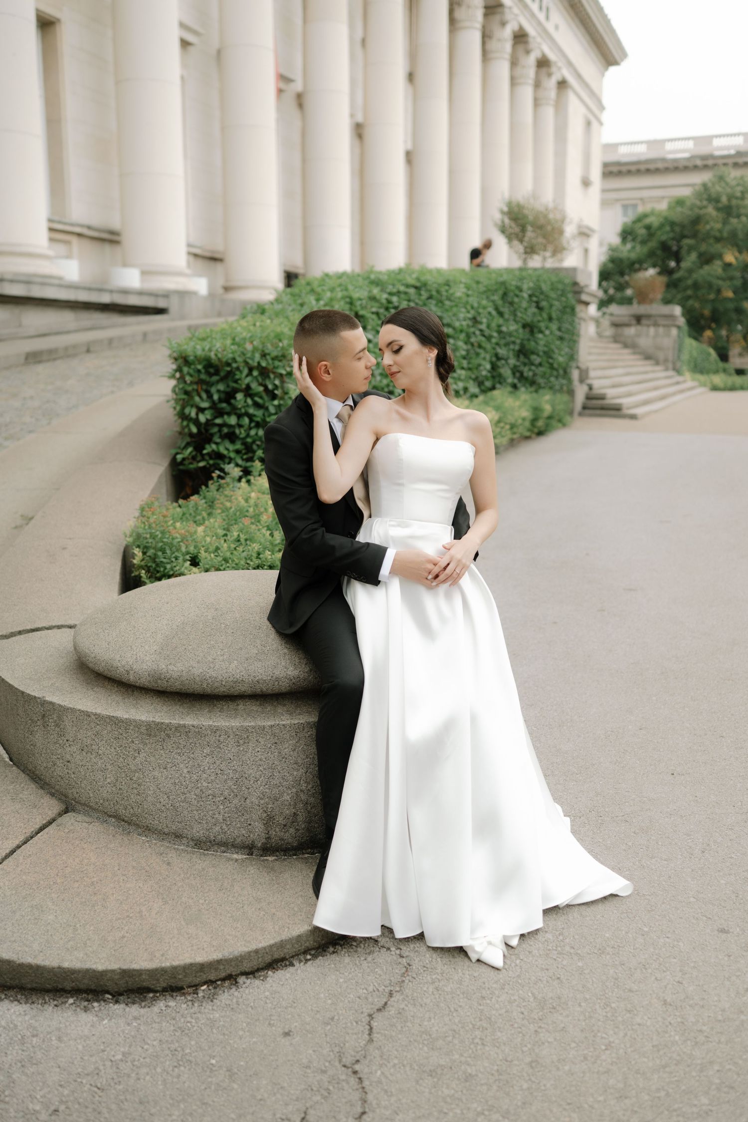 A couple in formal wedding attire share romantic moments on curved stone steps outside a classical building.