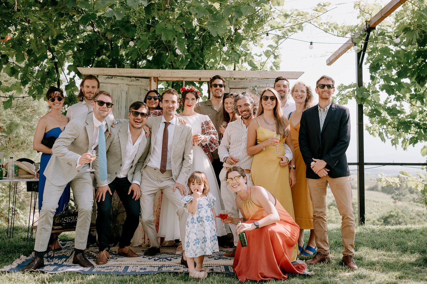 A group of wedding guests pose together outdoors under green trees on a sunny day.