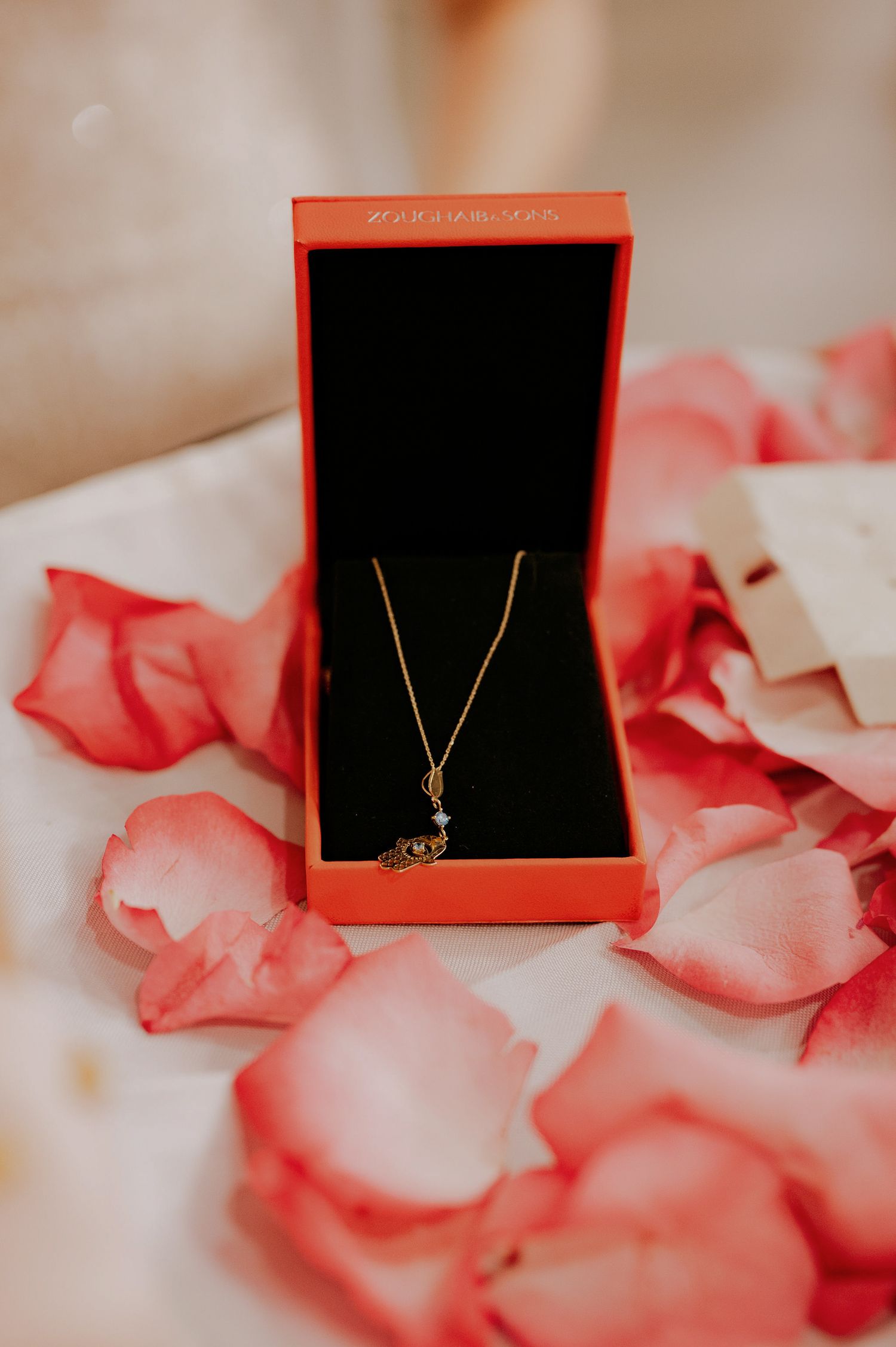Gold pendant necklace in red box surrounded by scattered pink and white rose petals.