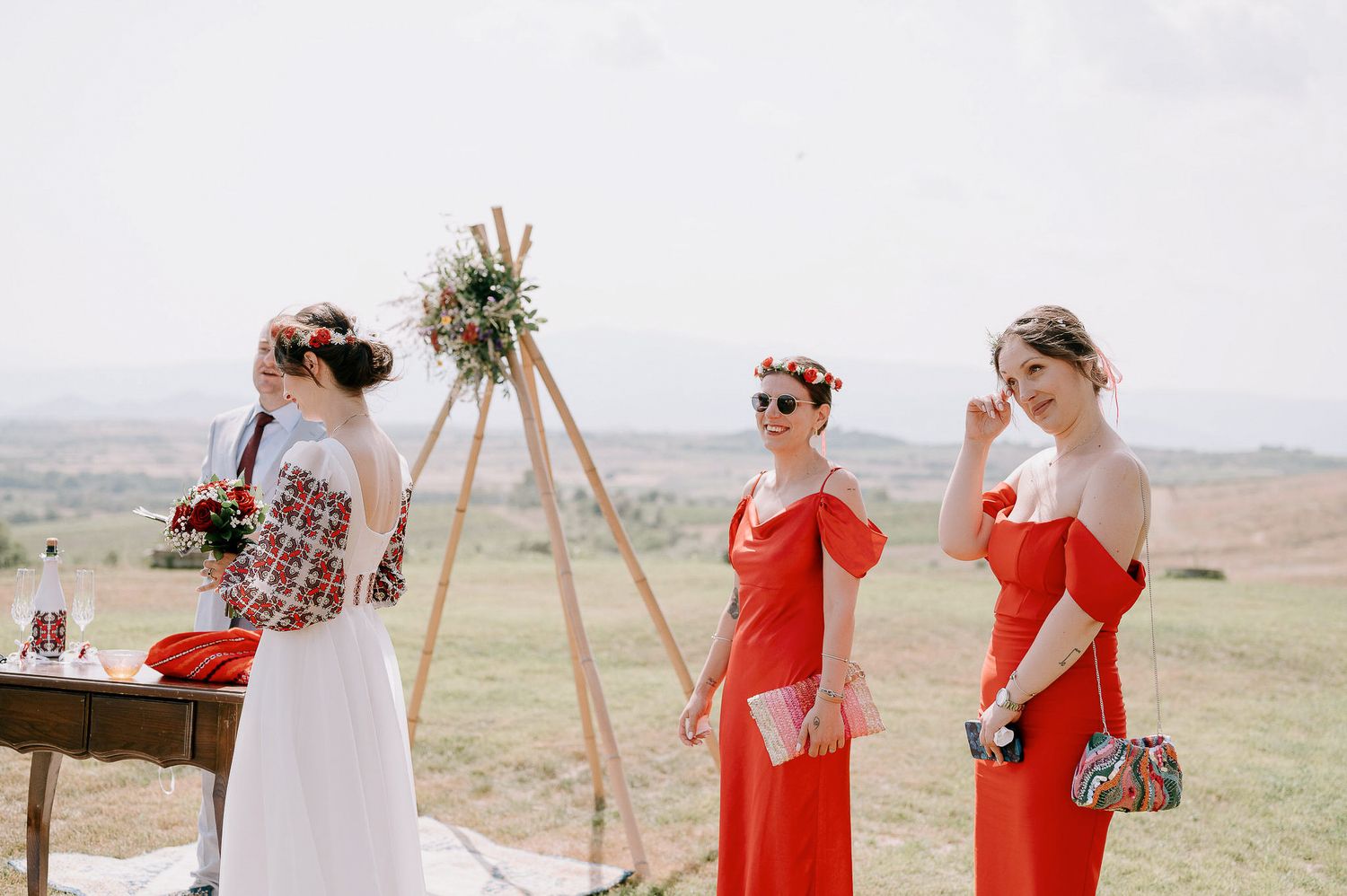Wedding guests gather on a grassy hillside with wooden decor and red accents at an outdoor ceremony.