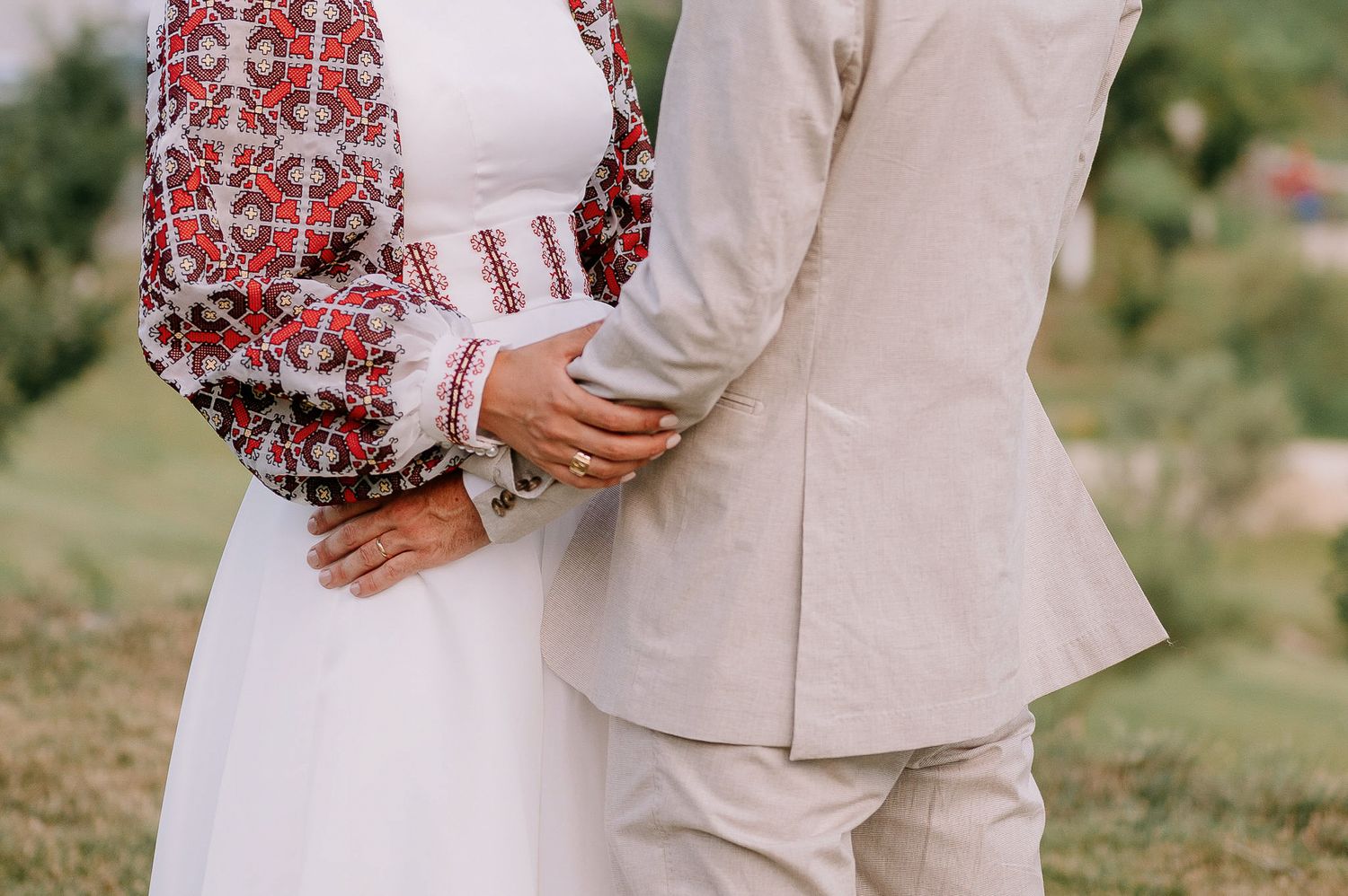 Romantic wedding couple embraces outdoors with mountains in background wearing traditional dress with red embroidery details.