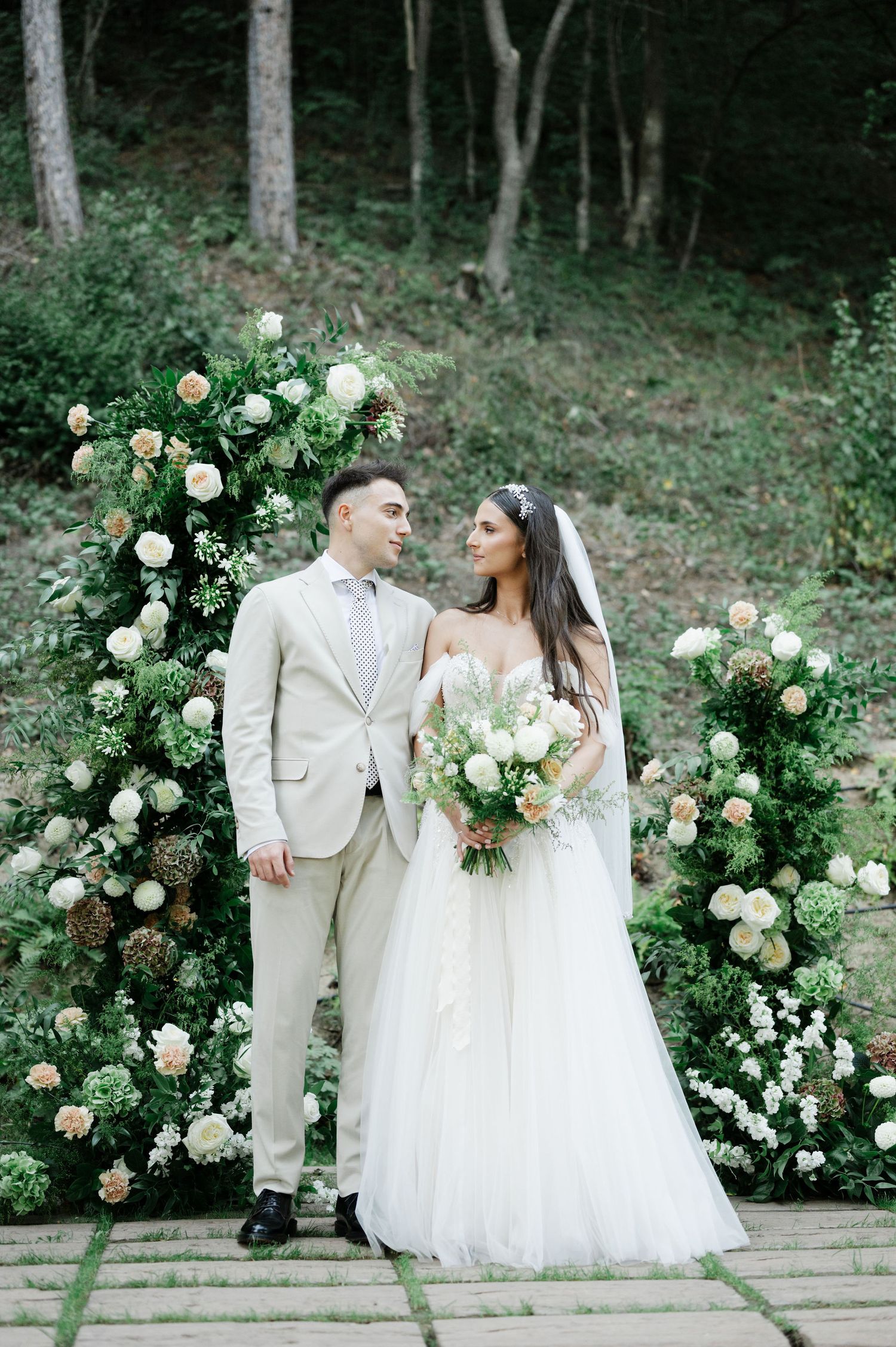 Elegant outdoor wedding ceremony setup with lush rose arch and stone pathway in a wooded setting.