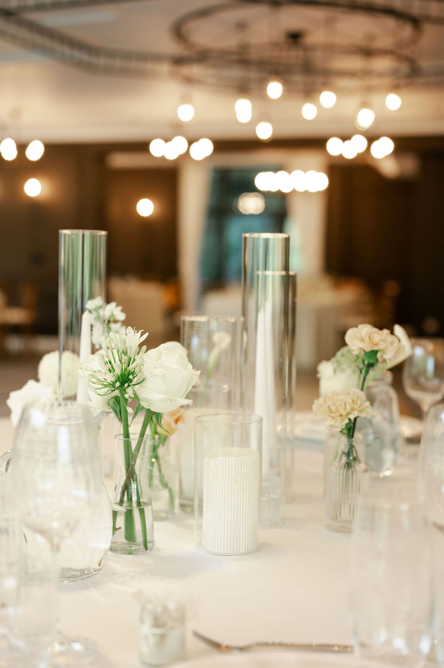 Close-up of minimalist wedding centerpiece featuring white flowers in glass vases and candles on white tablecloth.