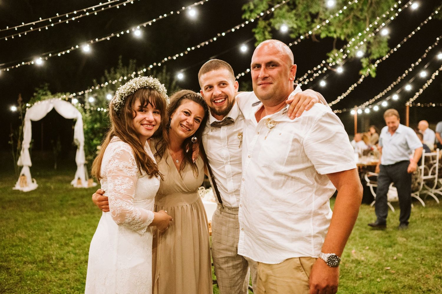 Wedding reception guests gather under romantic string lights at an outdoor evening celebration.