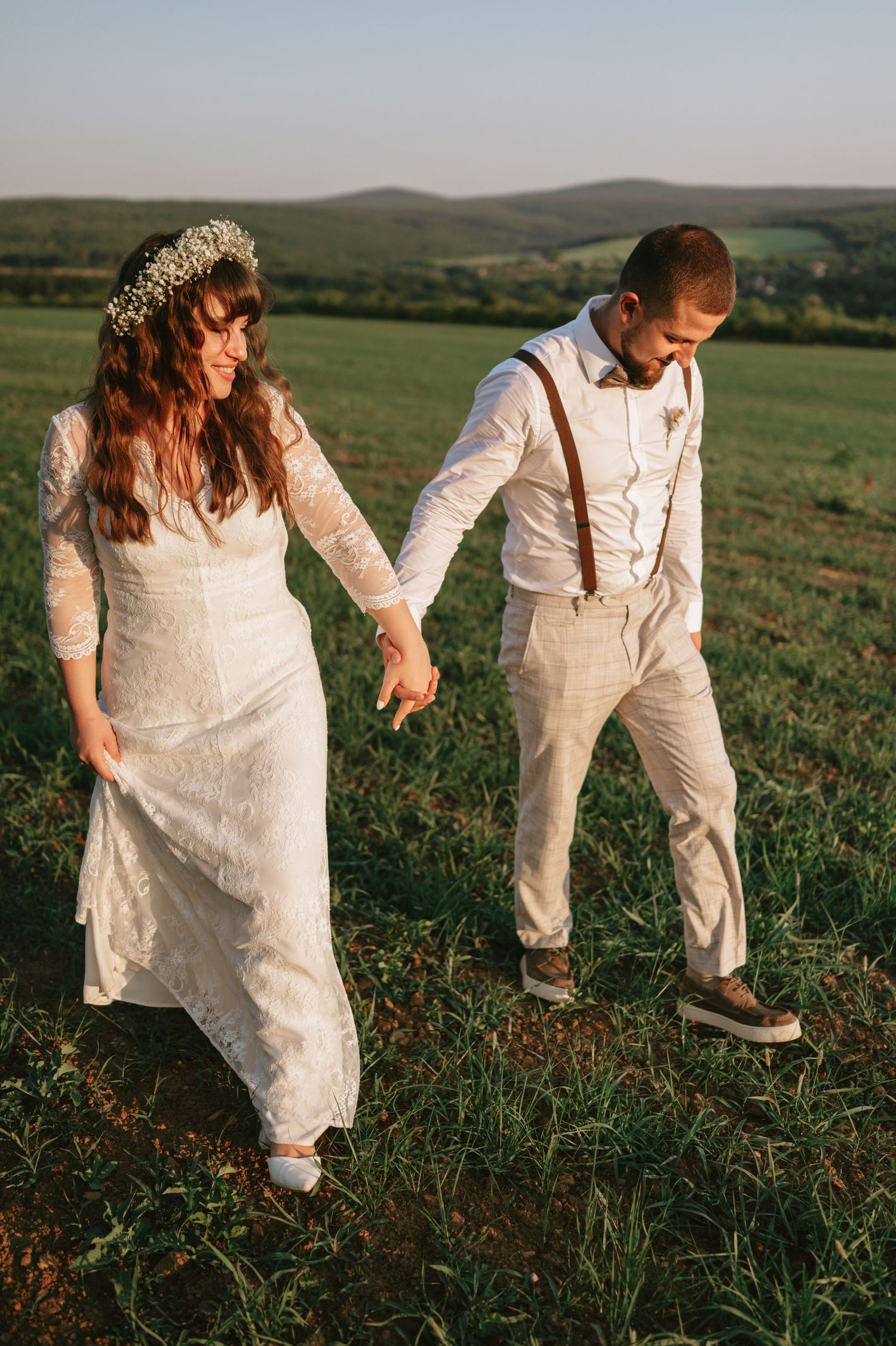 A couple in white outfits walks through a scenic green hillside at sunset.