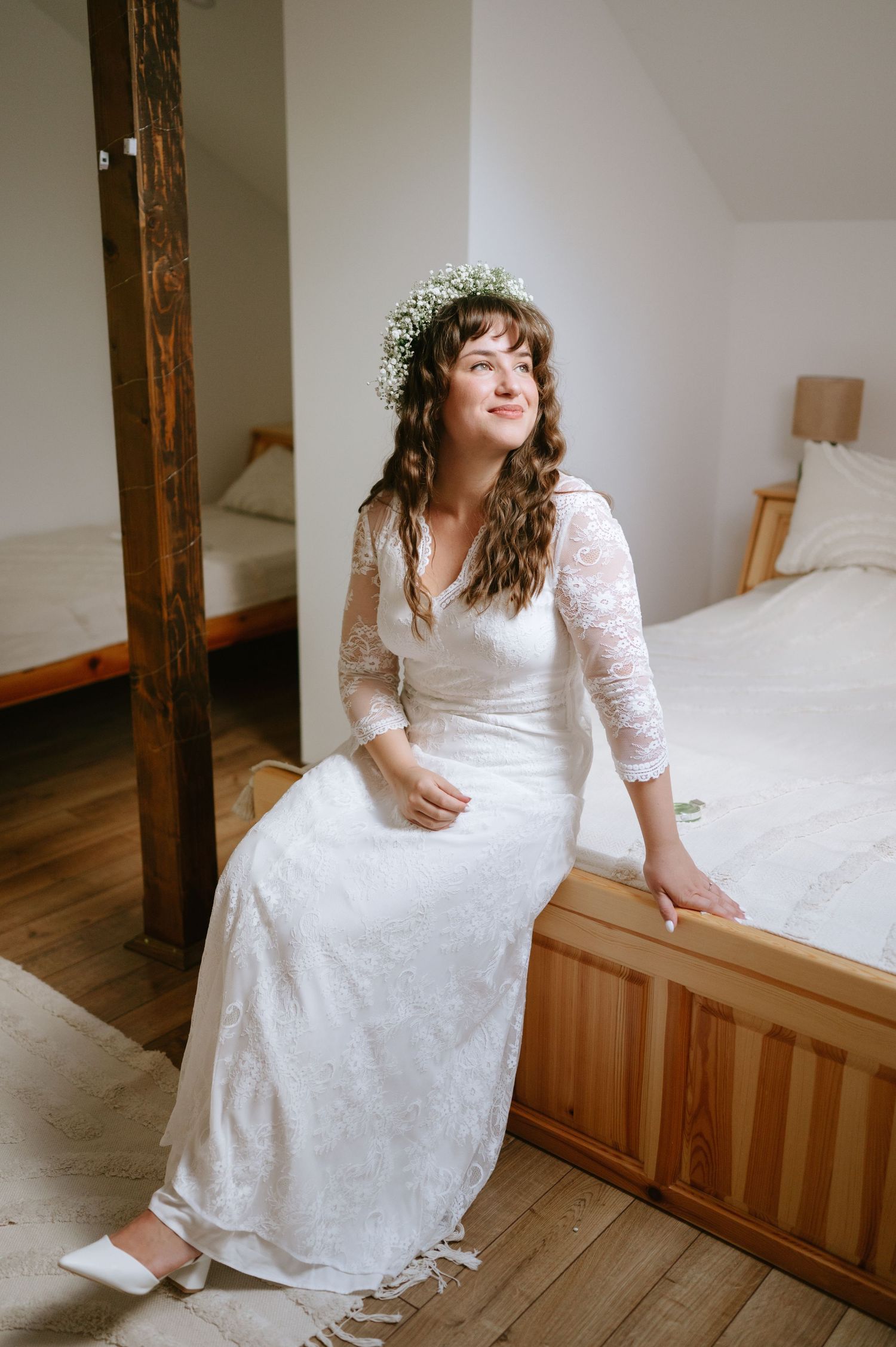 A white lace wedding dress with long sleeves is photographed in a rustic indoor setting with wooden beams.