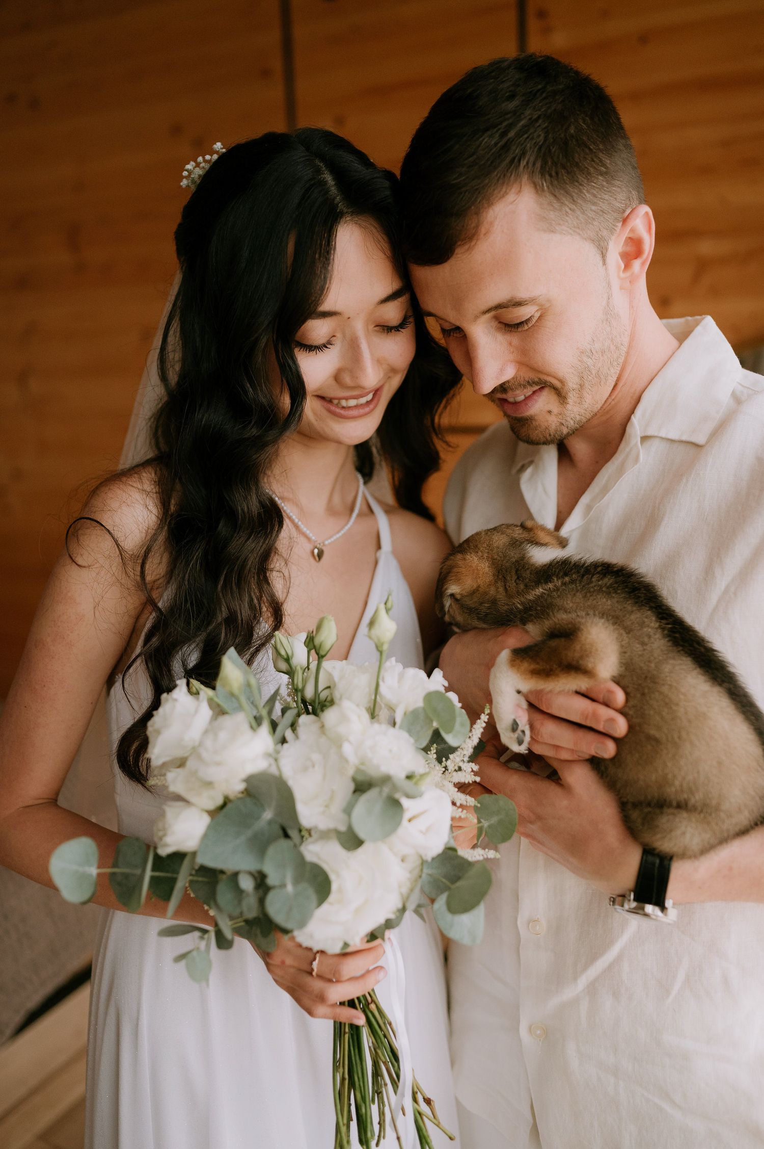 A couple shares tender moments with their dog while holding a white bouquet in a wooden room.