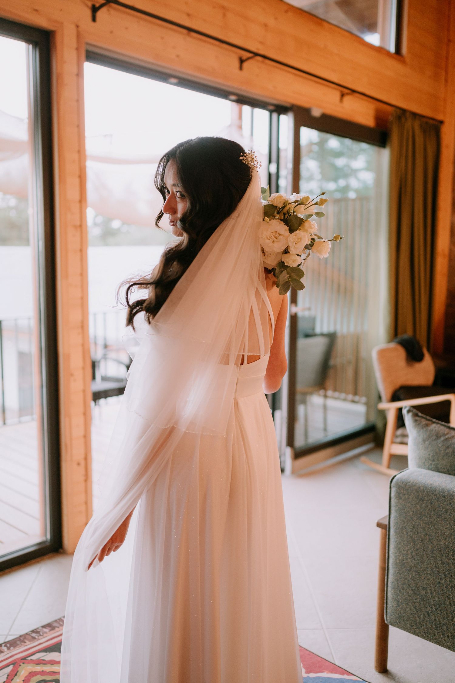 Bride in flowing white dress and veil standing by large windows in wooden interior.