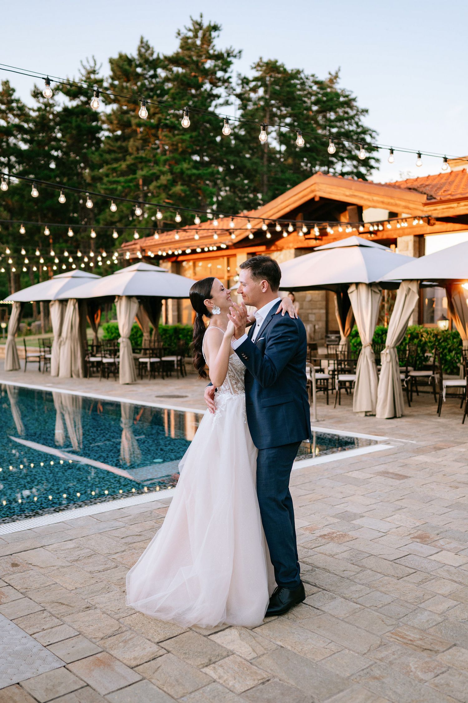 Wedding couple dancing by a poolside venue with string lights and white cabanas in the background.
