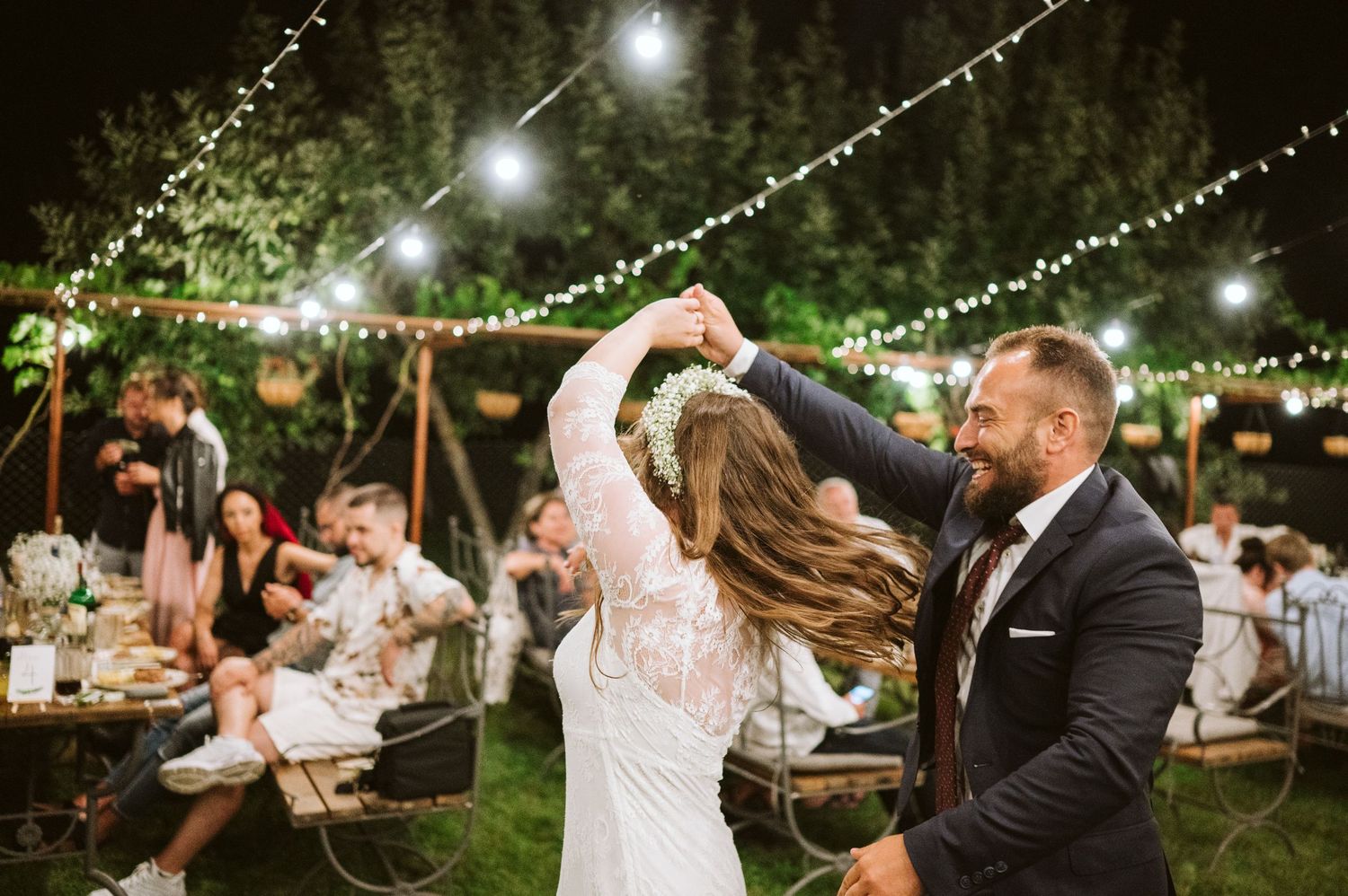 Wedding reception guests dancing and celebrating under romantic string lights in an outdoor garden setting.
