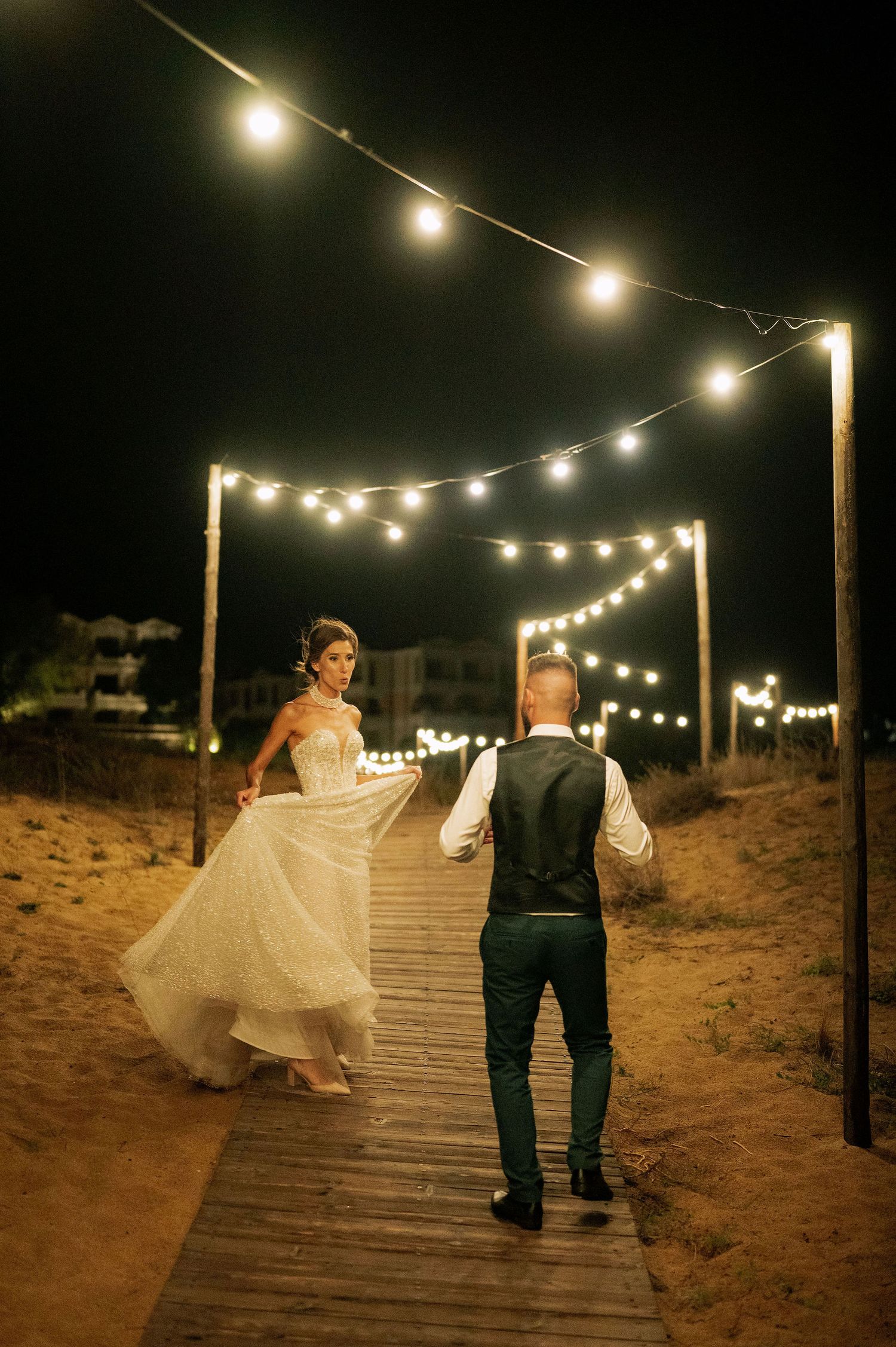 A bride in a sparkly gown twirls on a wooden walkway under string lights at night.