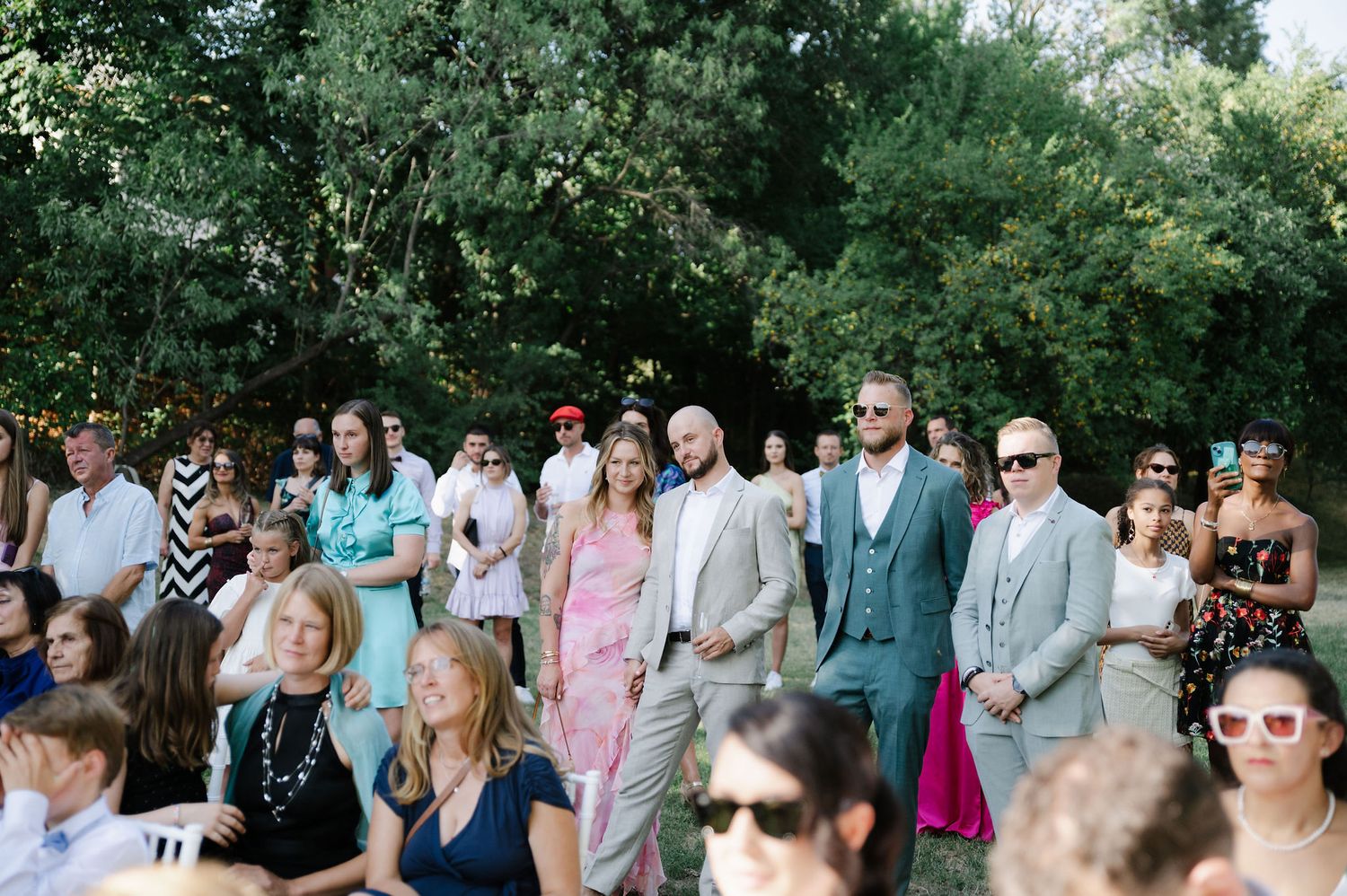 Wedding guests in colorful attire gather outdoors for a sunny celebration.