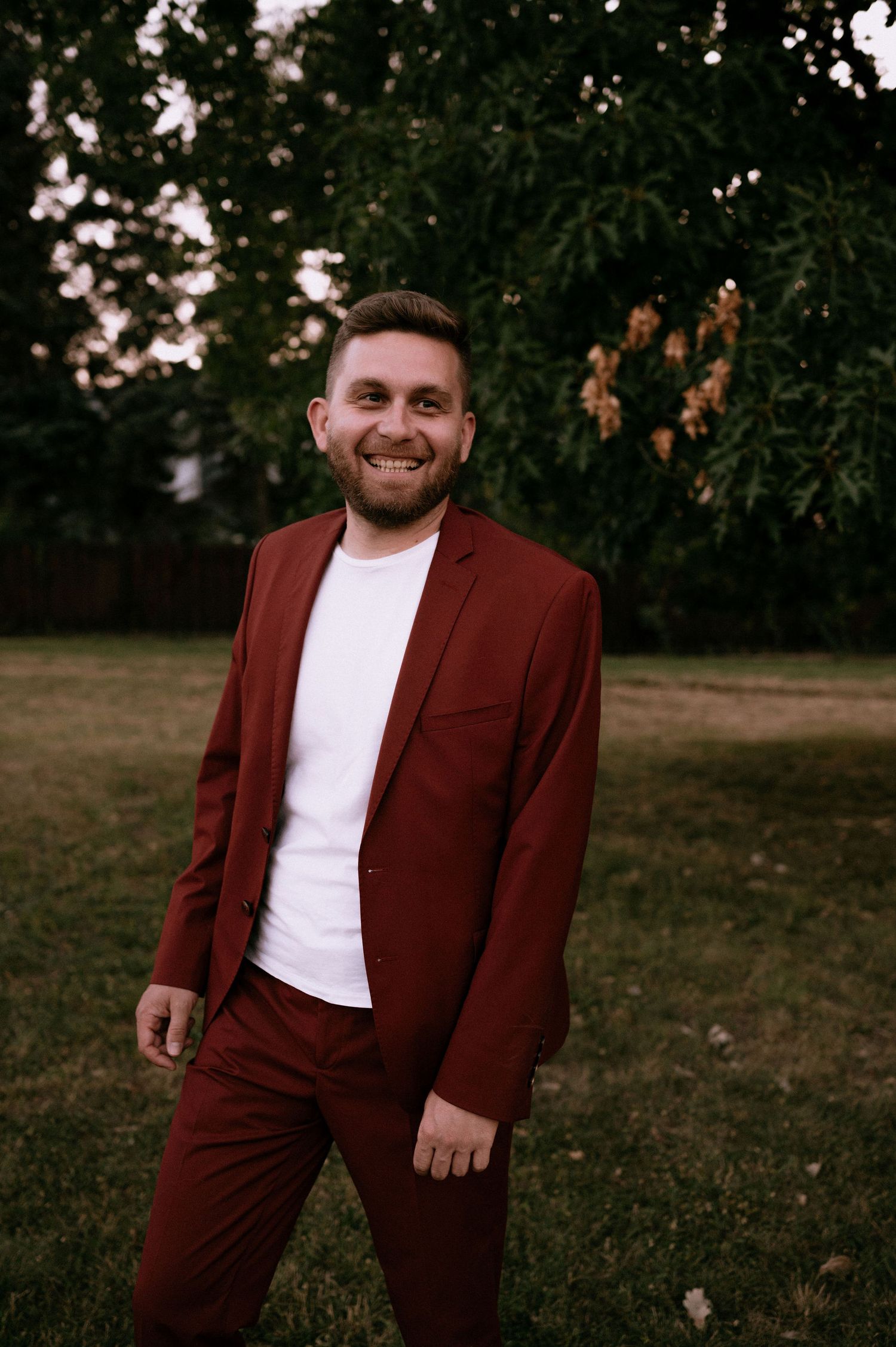 Person in burgundy suit and white shirt poses outdoors during golden hour.