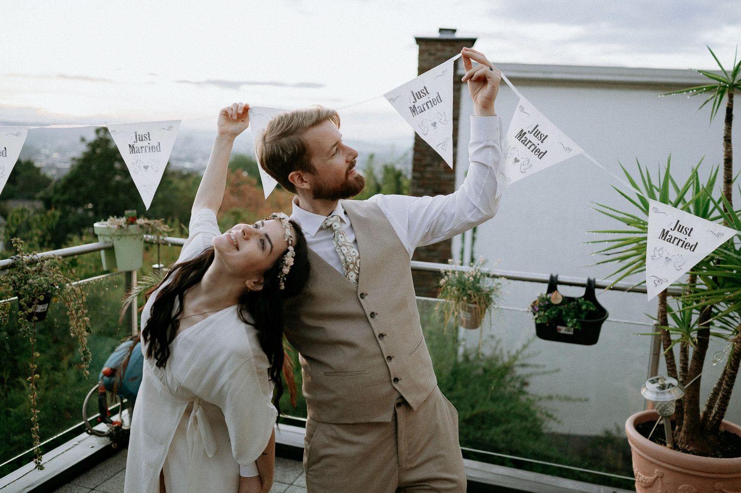 Wedding couple stands on a balcony with decorative white banners and potted plants.