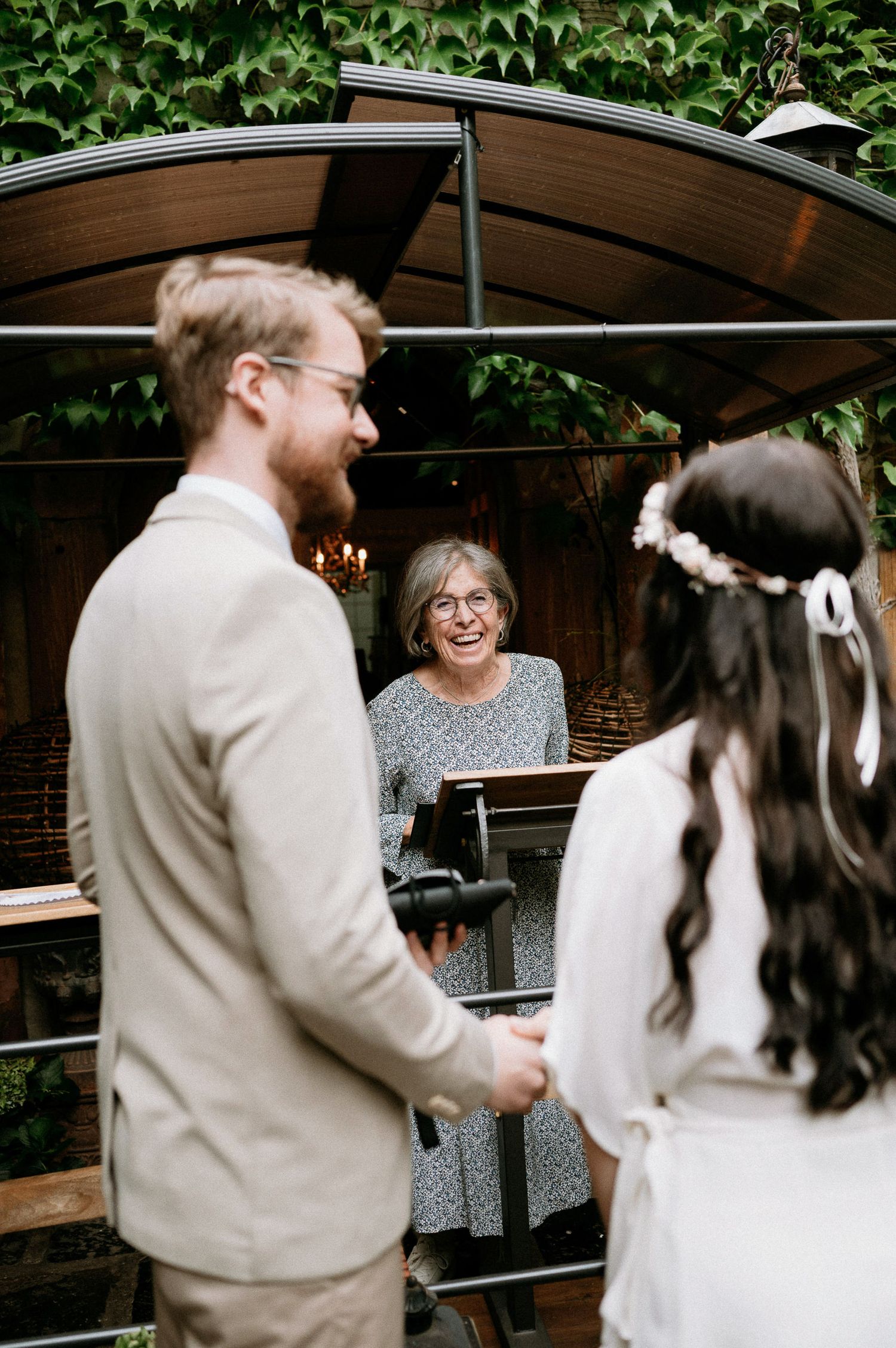 A wedding ceremony takes place under a wooden canopy covered in greenery.