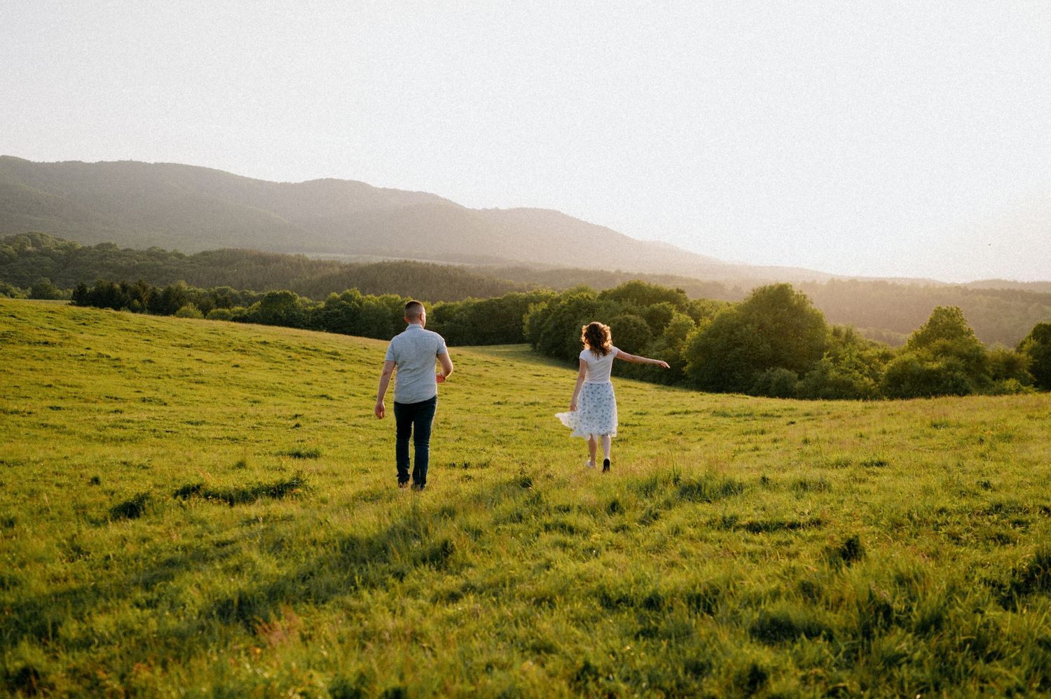 Couple walking hand in hand through a scenic hilltop meadow at sunset.