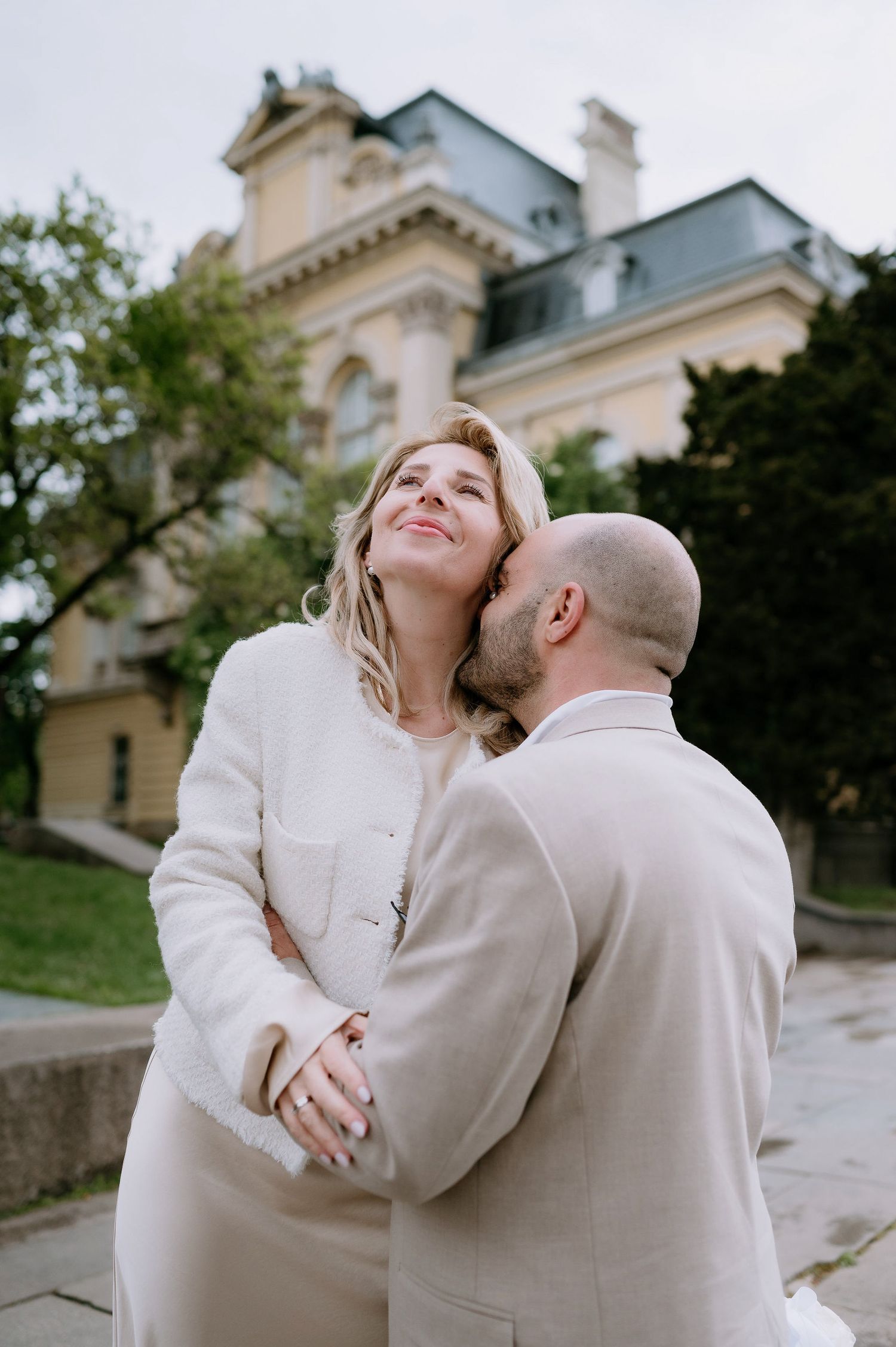 Two people in white clothing share a loving moment outside an elegant architectural backdrop.