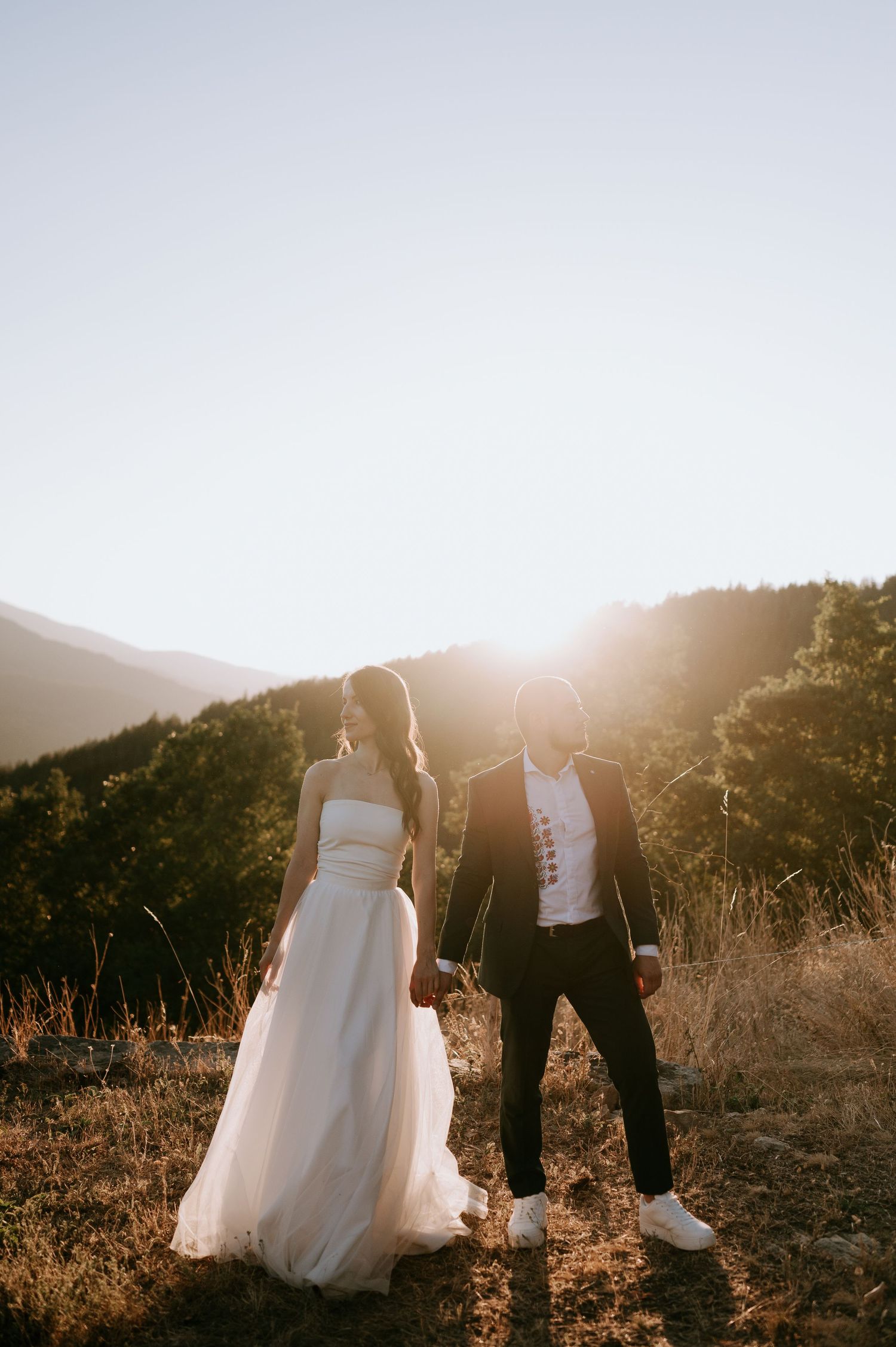 A couple in wedding attire walks together during golden hour in the mountains.