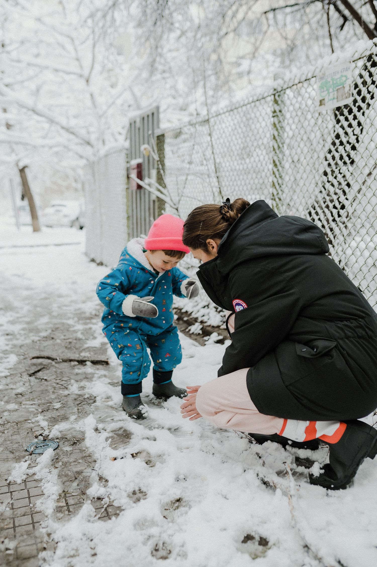 A small child in a blue snowsuit explores fresh snow in a winter scene while an adult crouches nearby.