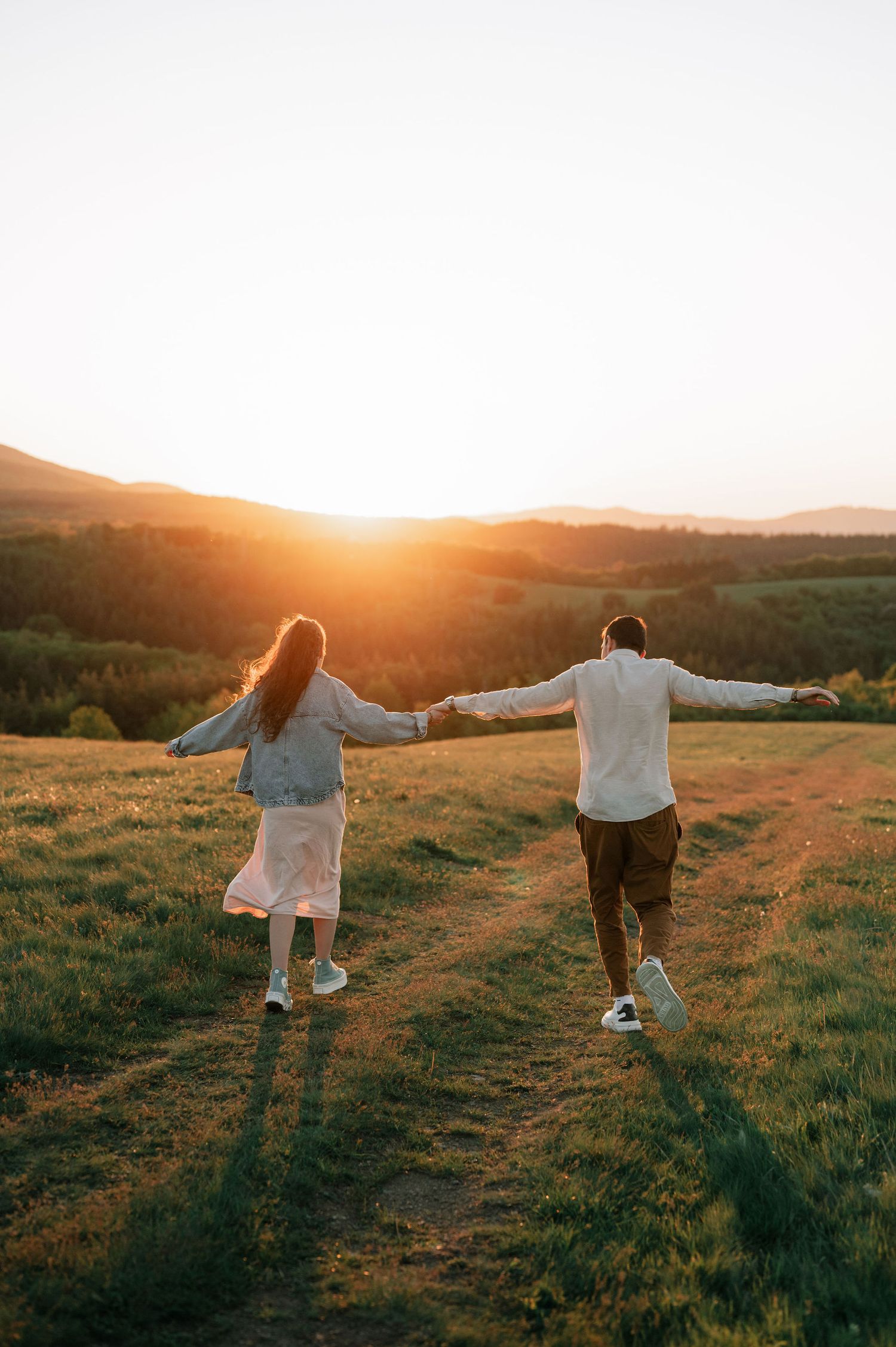 Two people with outstretched arms walk through a grassy hillside during a golden sunset.
