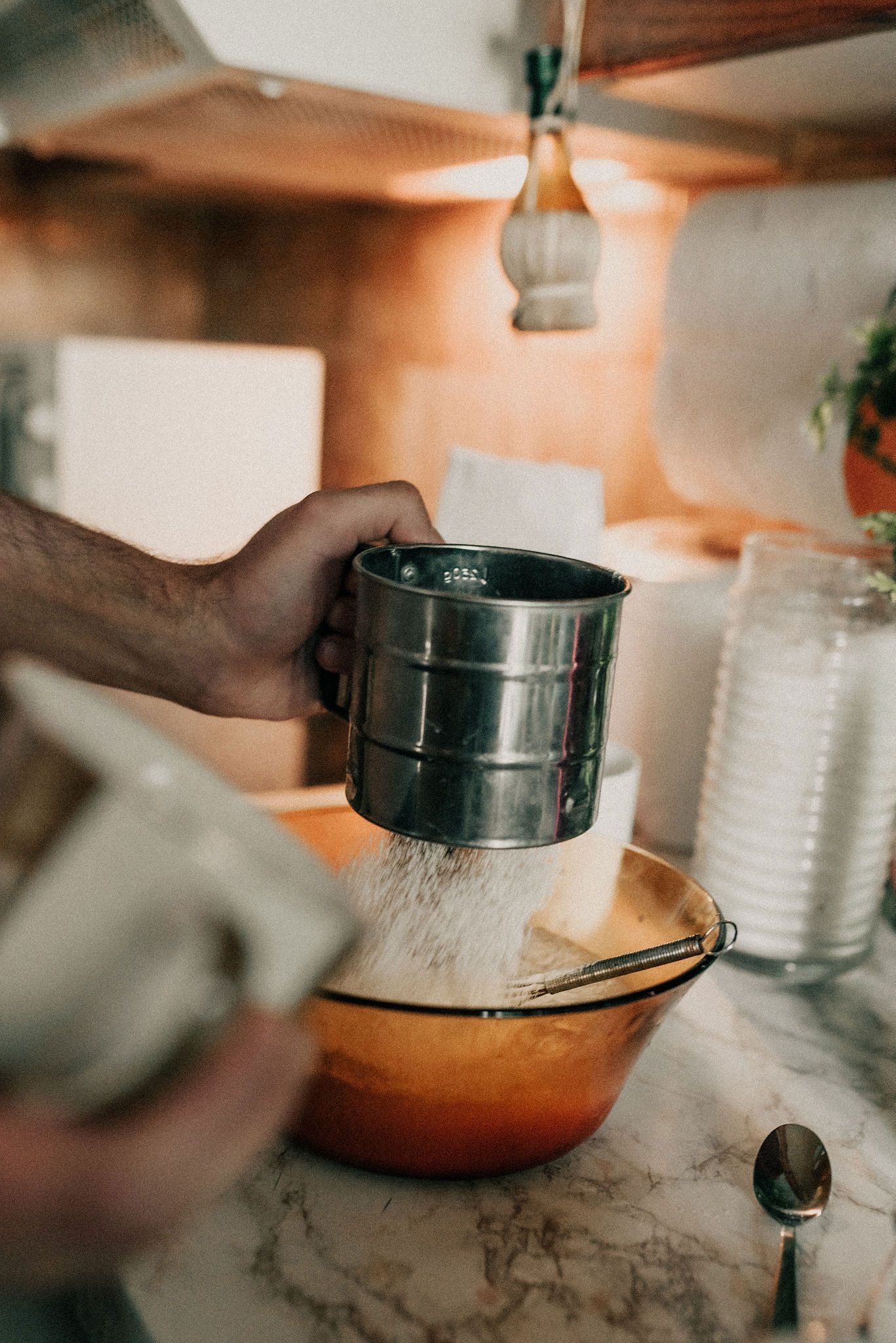 Close-up shots of hands sifting flour through a metal sieve into a mixing bowl on a marble countertop.