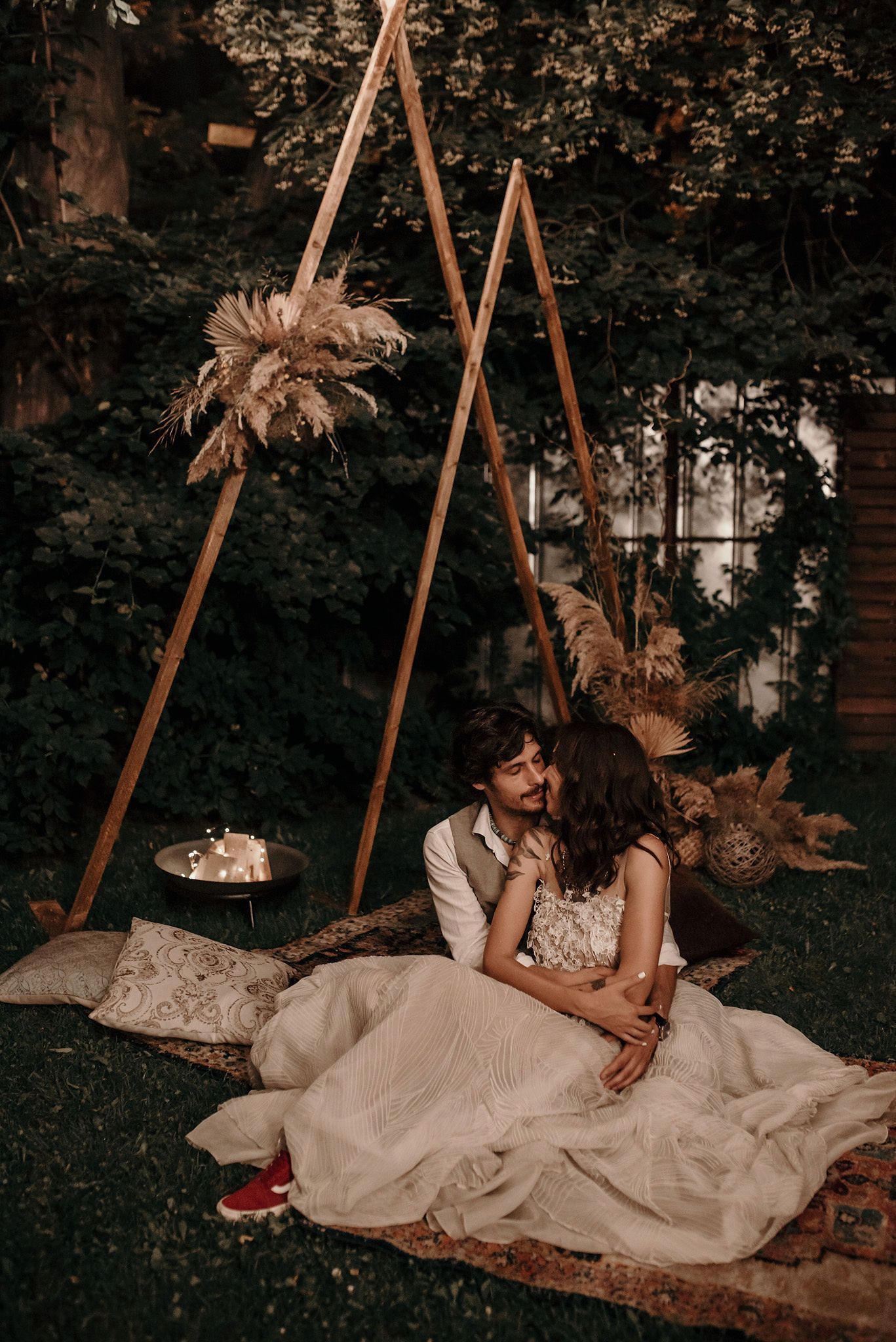 Romantic outdoor wedding setup with triangular wooden frames decorated with pampas grass and a cozy blanket seating area.
