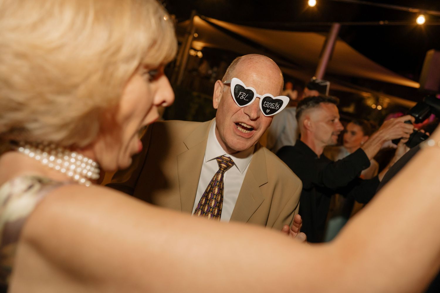 Bald man wearing heart-shaped novelty sunglasses dancing and laughing at a lively indoor party event.