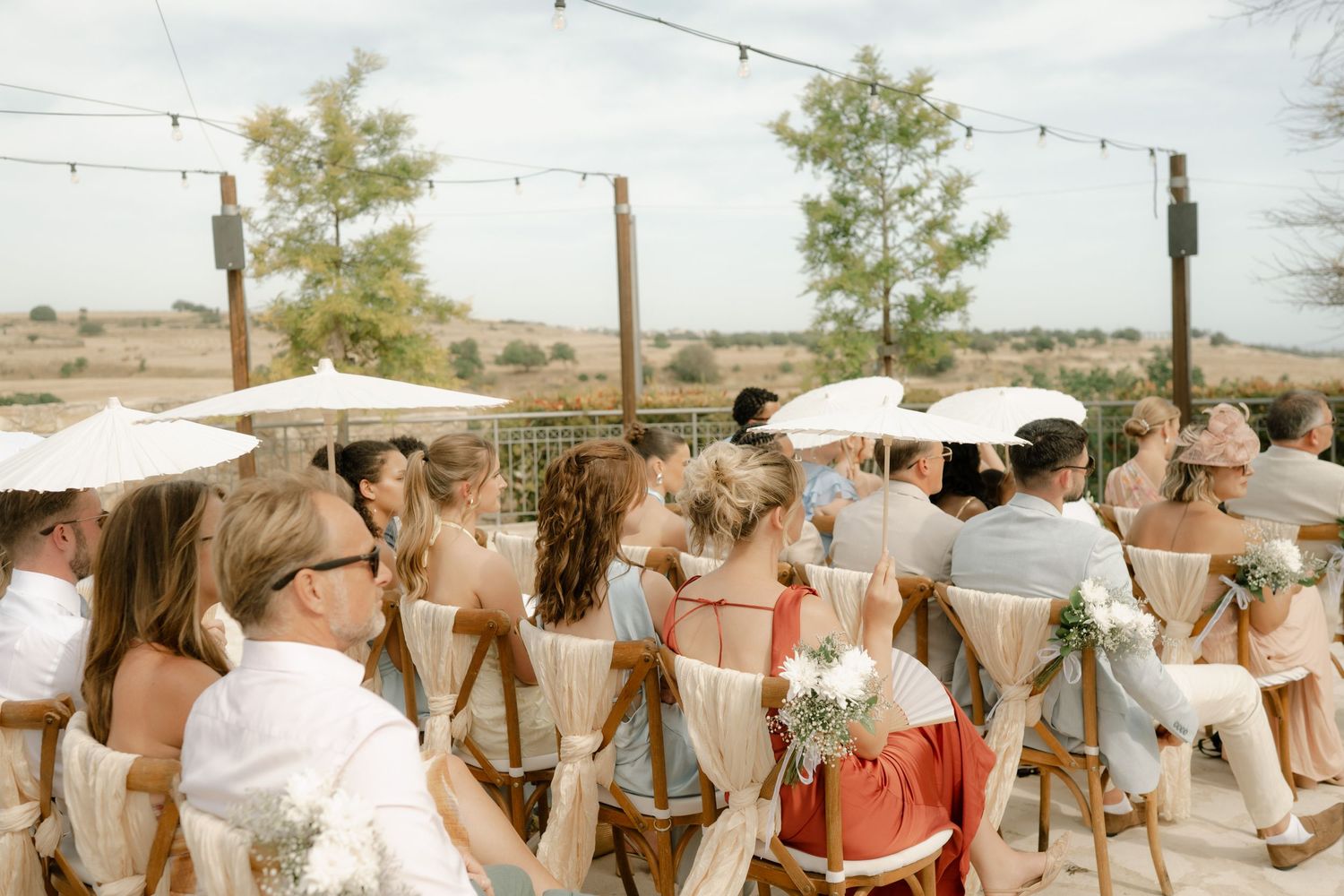 Outdoor wedding ceremony with guests seated on wooden chairs holding white parasols in a desert landscape.