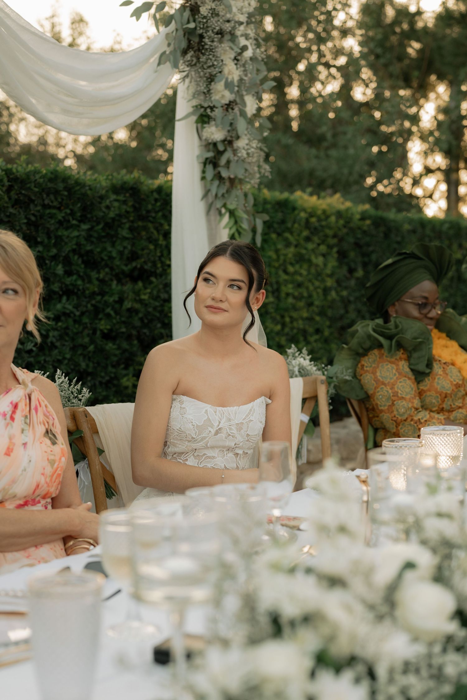 Bride in strapless lace gown seated at outdoor wedding reception table with floral decor and candles.