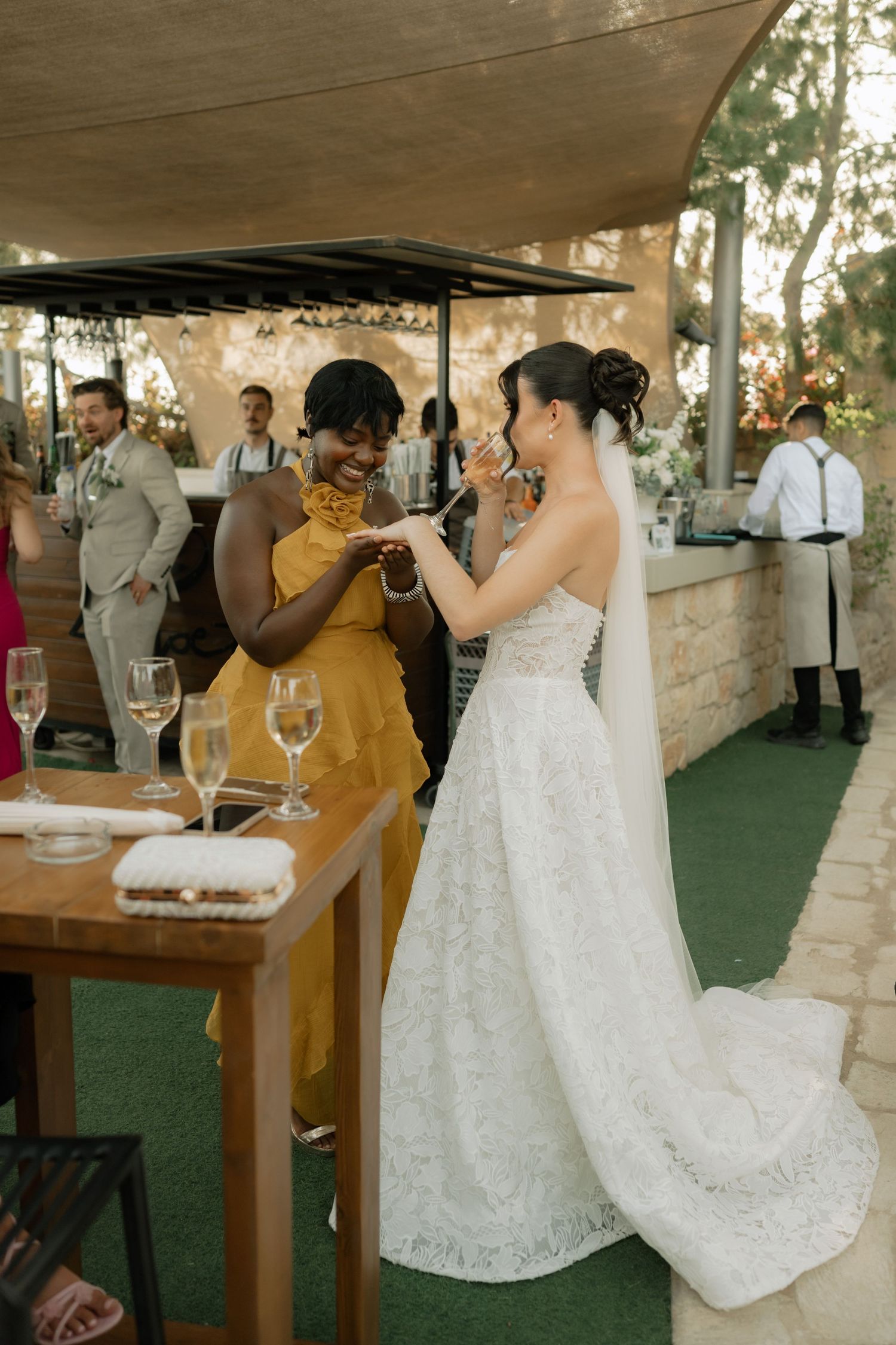 Bride in white lace gown and veil clinking glasses with smiling guest in yellow dress at wedding.