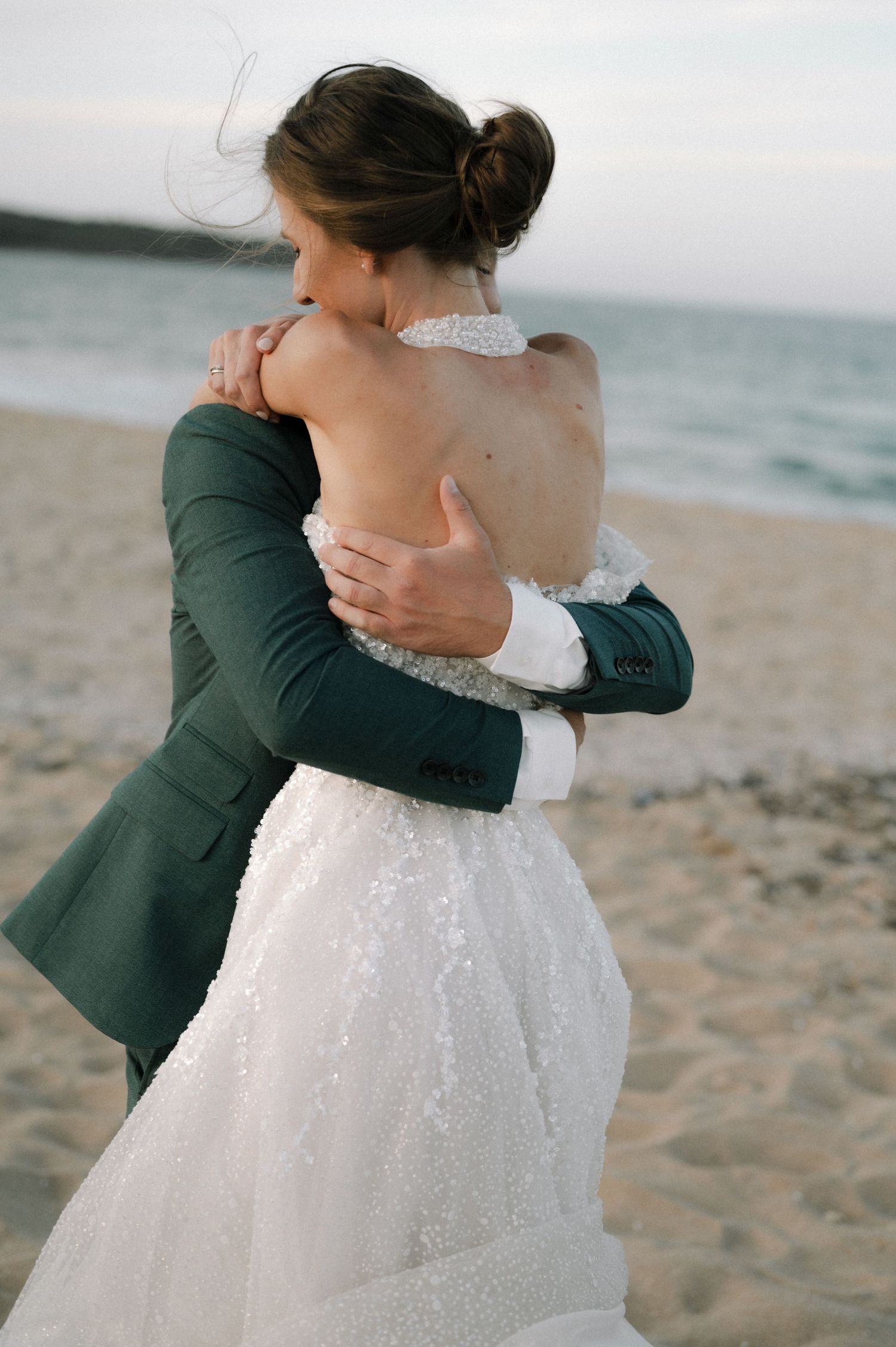 Bride and groom embracing on sandy beach, bride in backless lace gown, groom in dark green suit at sunset.