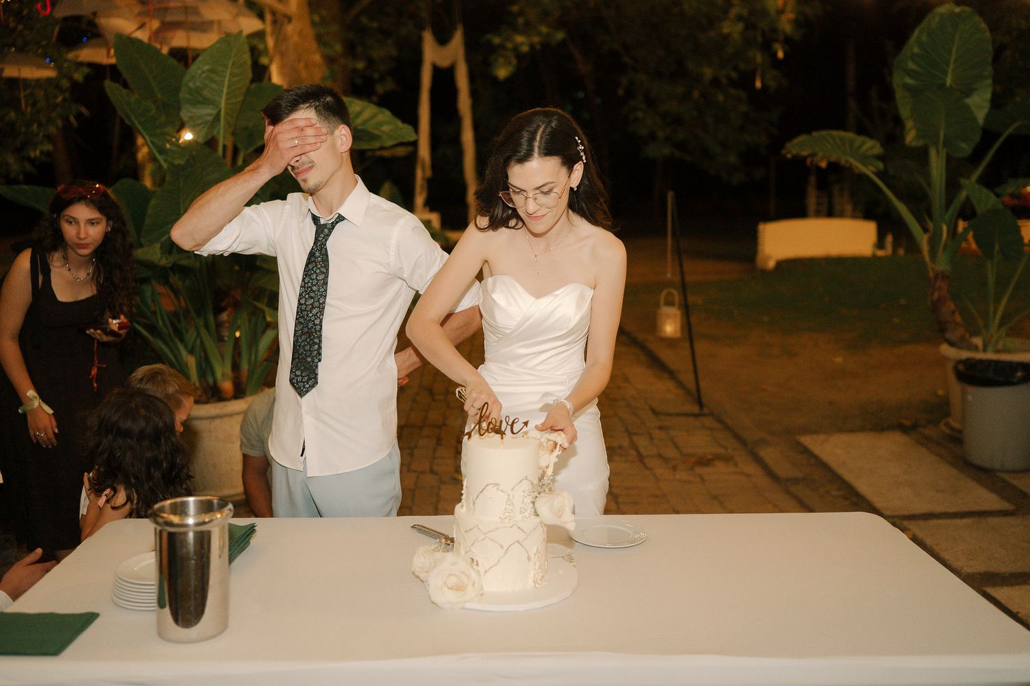 A couple cuts into their elegant white wedding cake during an evening outdoor reception celebration.